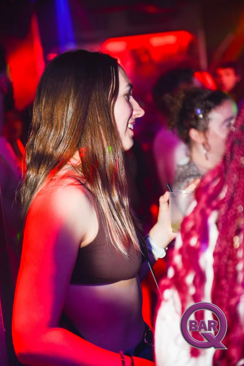A woman with long brown hair holding a drink in a nightclub, surrounded by other people, with neon and LED lighting creating a colorful atmosphere.