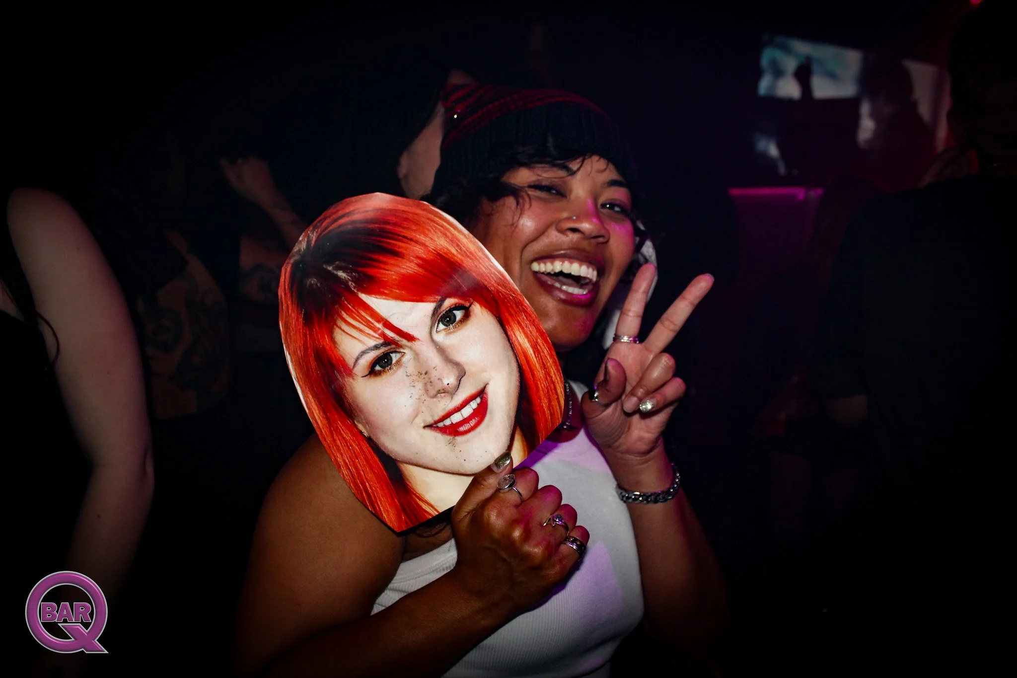People at a nightclub, one person holding a photo of a woman with red hair and freckles, making a peace sign, with others in the background.