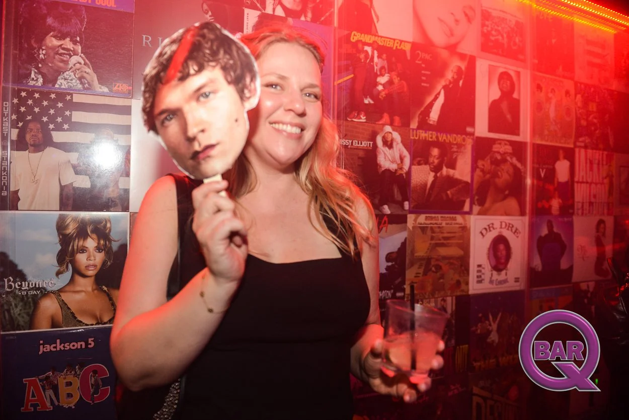 A woman with long blonde hair smiling while holding a cocktail glass in a karaoke bar. She is holding a large cutout mask of a man's face on a stick in front of her face. The background features a wall covered with posters of various music artists an