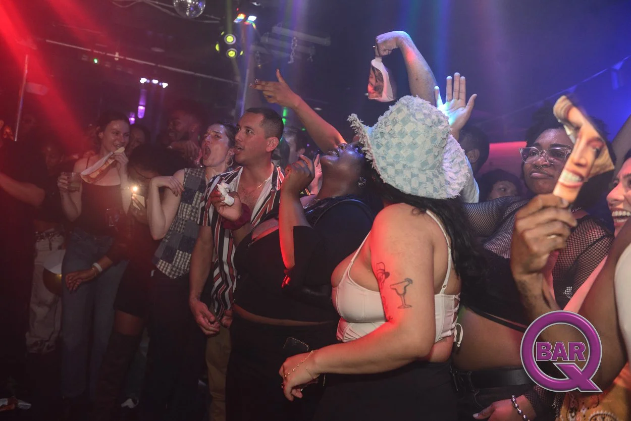 People dancing and having fun at a nightclub or party with colorful lighting, some holding drinks and masks. A woman wears a large sunhat and has a tattoo of a martini glass on her arm. The bottom right corner has a purple logo that reads 'BAR Q'.