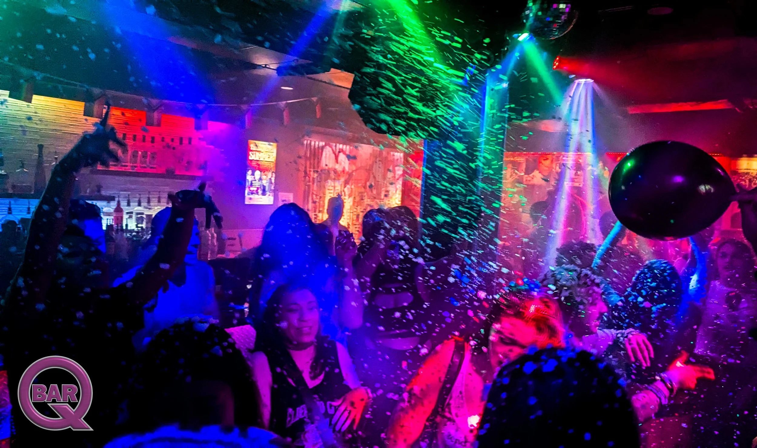 People dancing at a party with colorful laser lights, balloons, and confetti in a nightclub or bar setting.