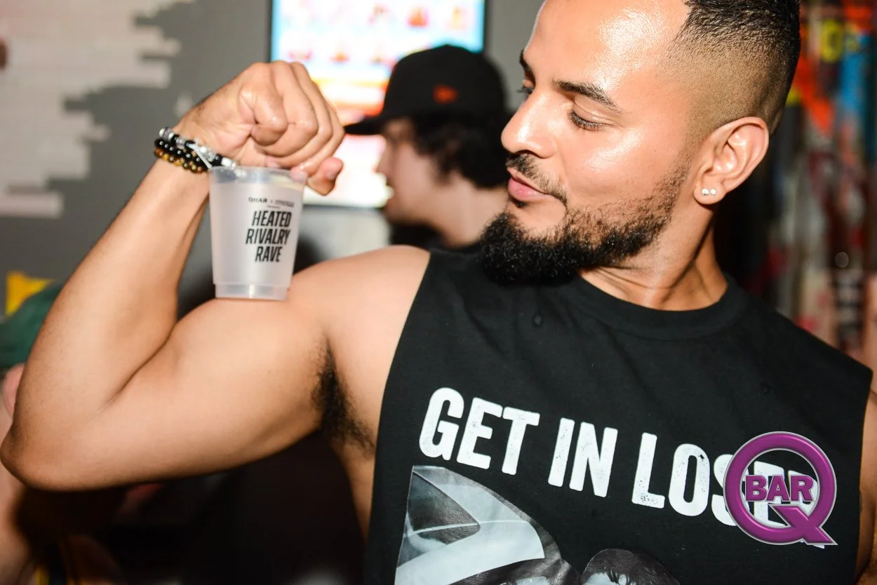 A man flexing his bicep and looking at it, wearing a sleeveless shirt with the text 'GET IN LOS' and a purple logo with the words 'BAR Q'. He is holding a cup labeled 'HEATED RIVALRY RAVE,' and has earrings and a bracelet. The background shows other 
