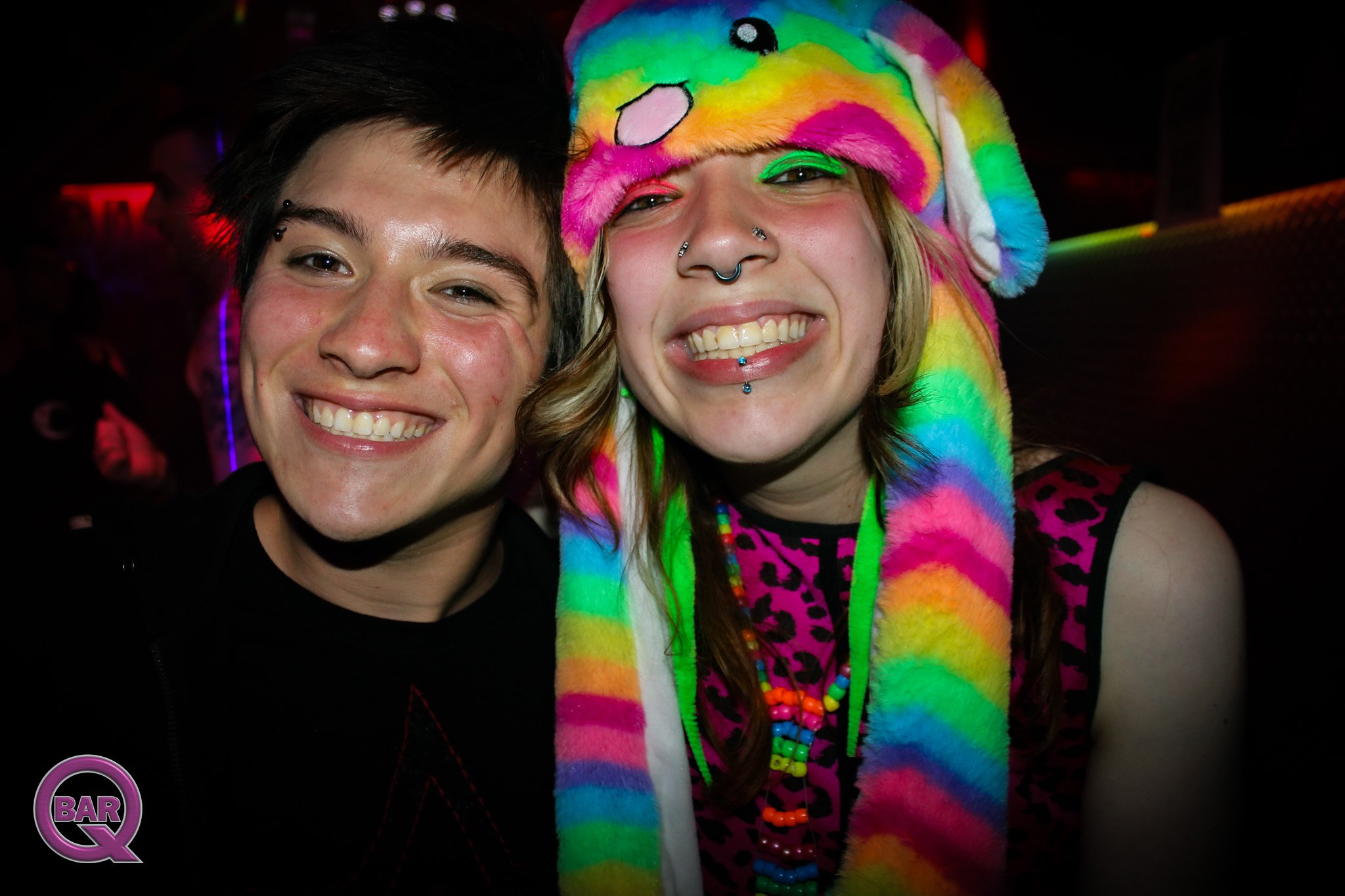 Two smiling individuals in a dark environment, one wearing colorful rainbow-themed clothing and accessories, including a plush rainbow hat with a cartoon face, bright makeup, and multiple necklaces, while the other has short dark hair and a pierced e