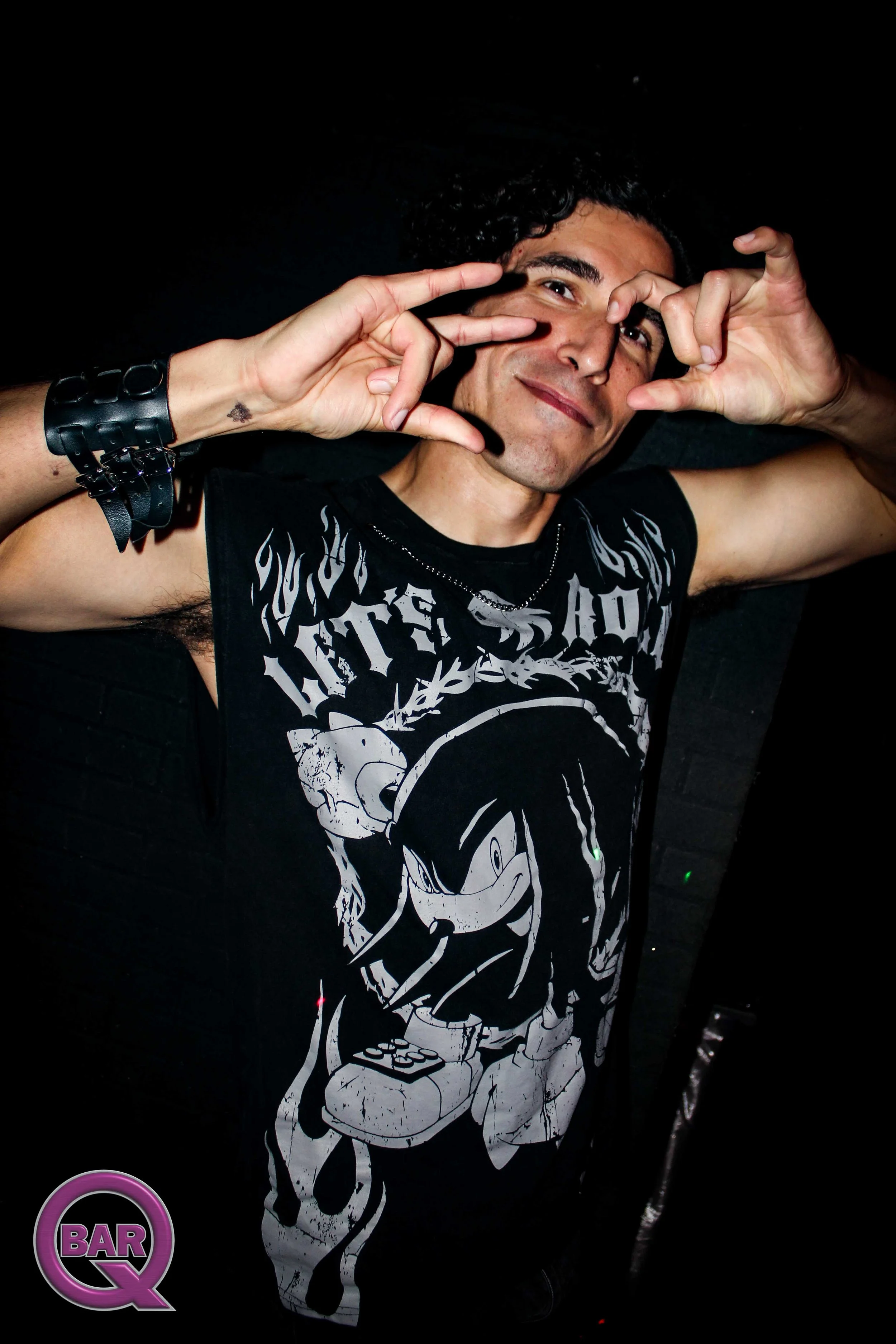 Person smiling and making a gesture with hands near face, wearing a black T-shirt featuring a cartoon character from Mickey Mouse and accessories, with a dark background and a purple circular bar Q logo at the bottom left corner.