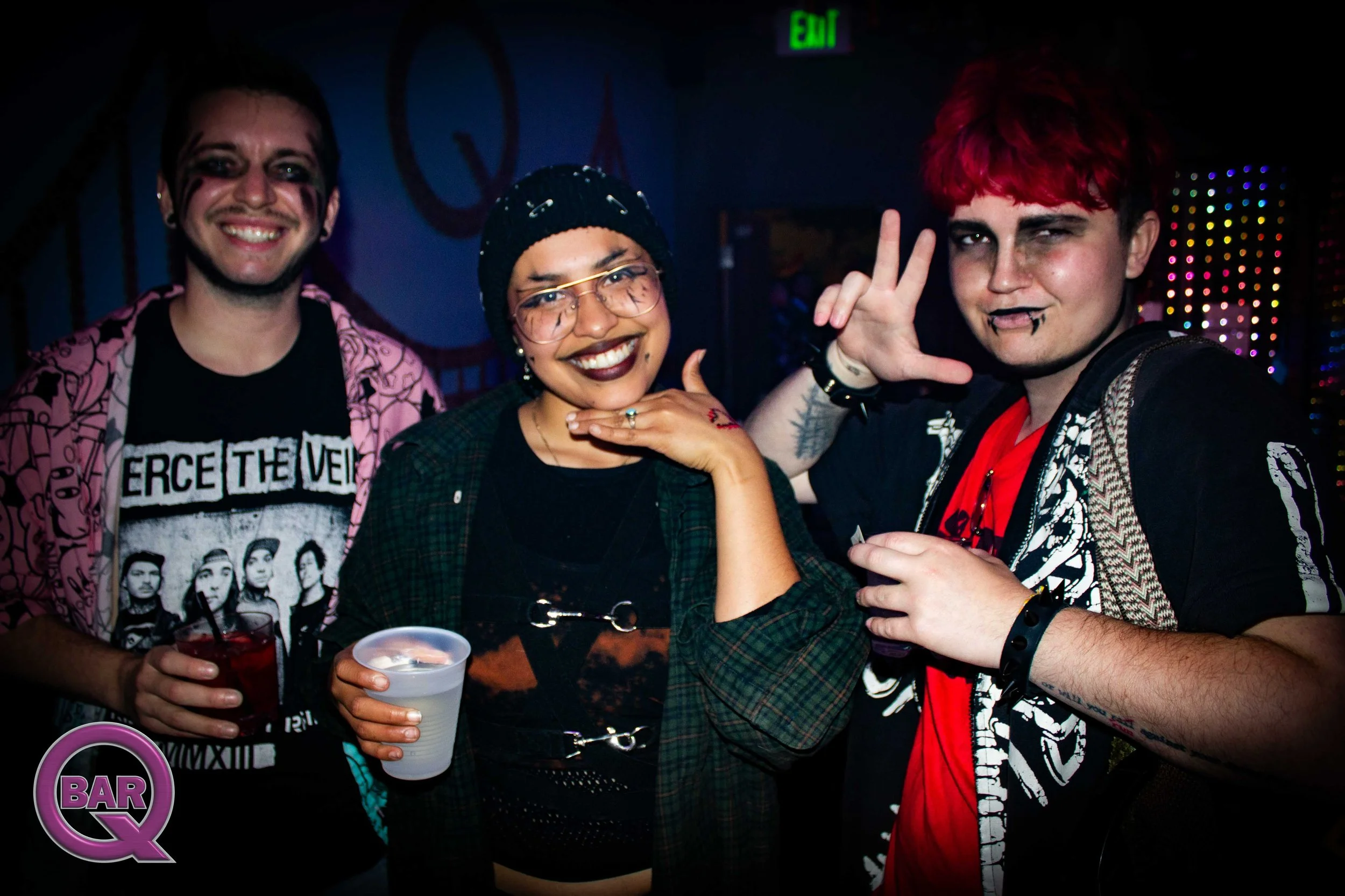 Three friends smiling and posing for a photo in a dark, colorful setting, possibly a nightclub or bar. The person on the left is wearing a pink patterned shirt and a black graphic T-shirt, holding drinks. The person in the middle is wearing glasses, 