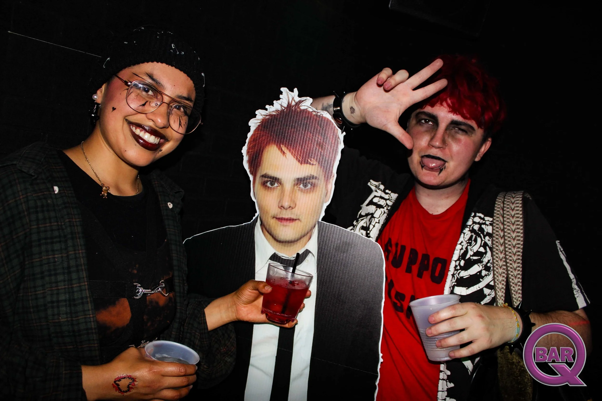 Two women and a cardboard cutout of a young man at a bar. The woman on the left is wearing glasses, a black beanie, and a dark patterned shirt, and is smiling. The woman on the right has vibrant red hair, is making a peace sign, sticking her tongue o