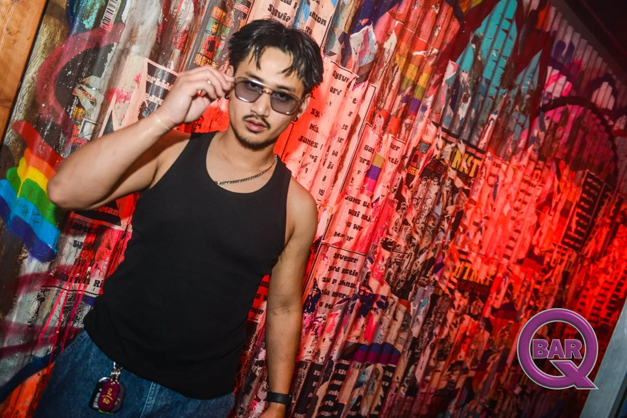 A young man with sunglasses, a black tank top, and jeans stands in front of a colorful graffiti wall. He is touching his sunglasses with his right hand, and has a keychain hanging from his pocket. There is a logo in the bottom right corner that says 
