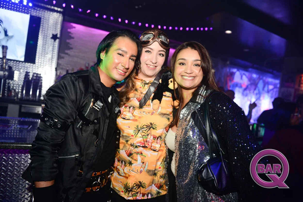 Three friends smiling in a nightclub with purple and pink lighting, standing in front of a bar, with a large screen and decorative lights in the background.