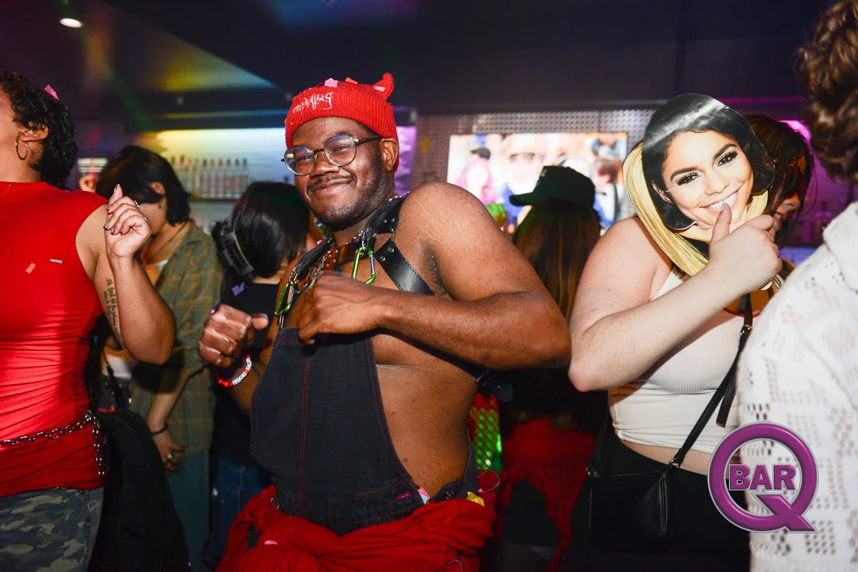 A group of people at a bar or nightclub, with a man at the center wearing a red hat and glasses, smiling and posing. A woman nearby holds a cutout microphone with a woman's face on it, giving a thumbs-up. The background has colorful lights and a scre