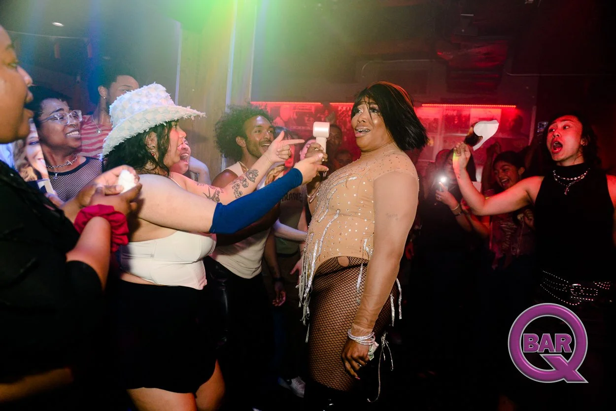 A group of people at a nightclub, with one woman in the center wearing a nude-colored top, fishnet stockings, and looking surprised or excited. Others are smiling and cheering around her, with colorful lights in the background and a "BAR Q" logo in t