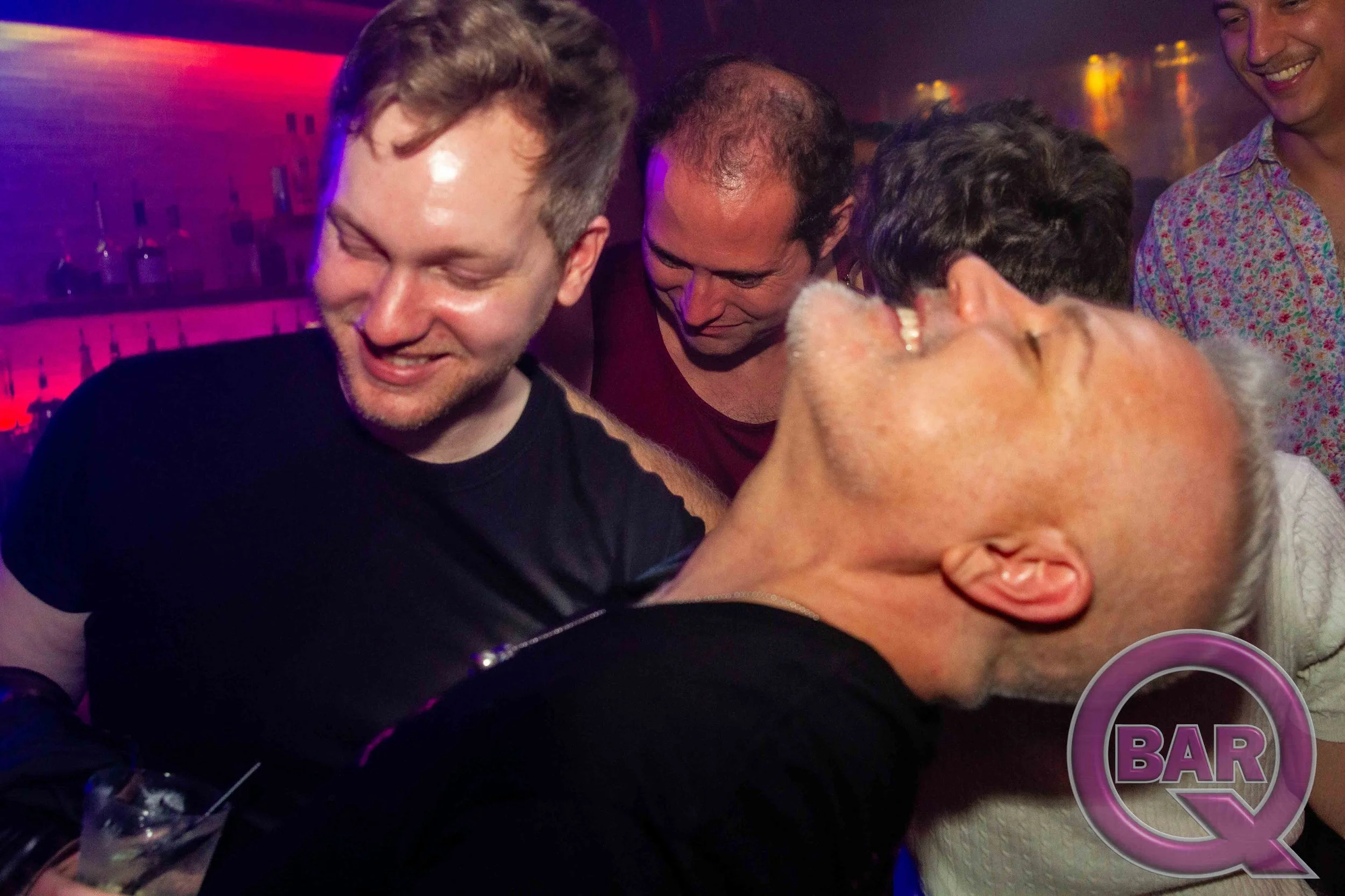 A group of people at a bar or nightclub, smiling and laughing, with one person holding another person close. The background is colorful with neon lighting, and a sign that says 'BAR' can be seen in the bottom right corner.