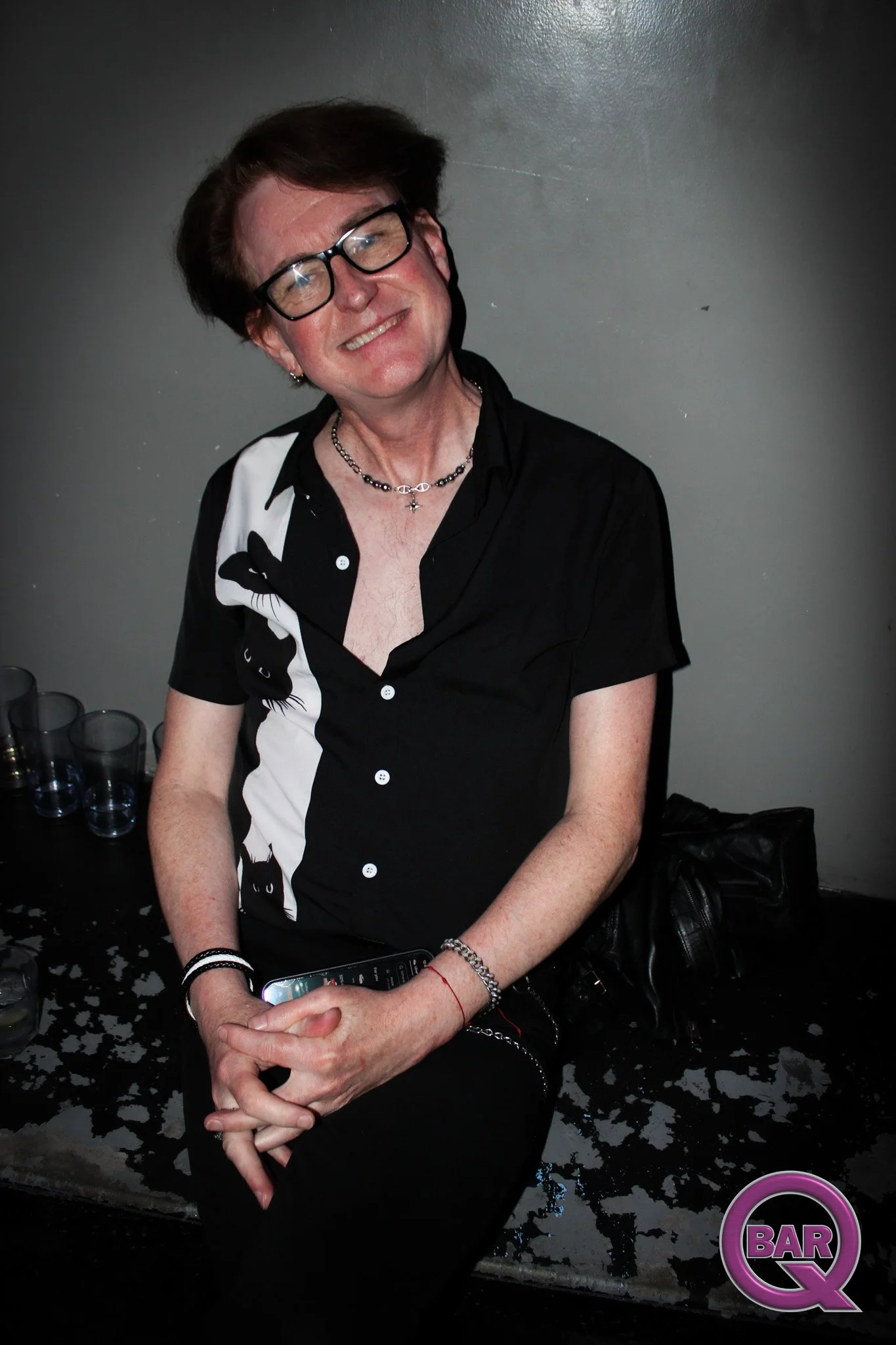 A smiling woman with short brown hair, wearing glasses, a black and white shirt, and a necklace, sitting at a bar with a dark wall background and drinks on the table.