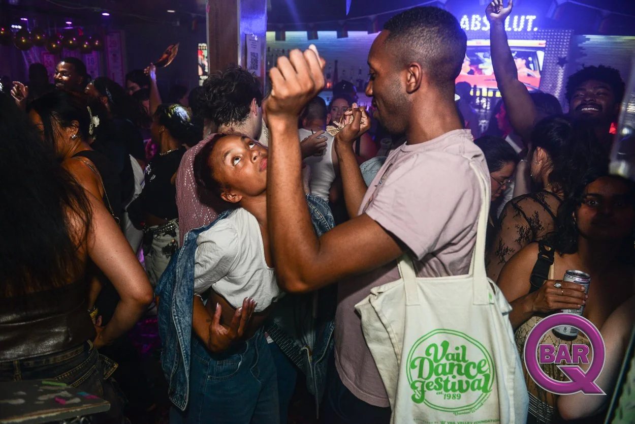 Crowd of people dancing at a nightclub, with a woman in a white shirt and jean jacket looking up at a man with a tote bag that reads 'Vail Dance Festival 1989'.