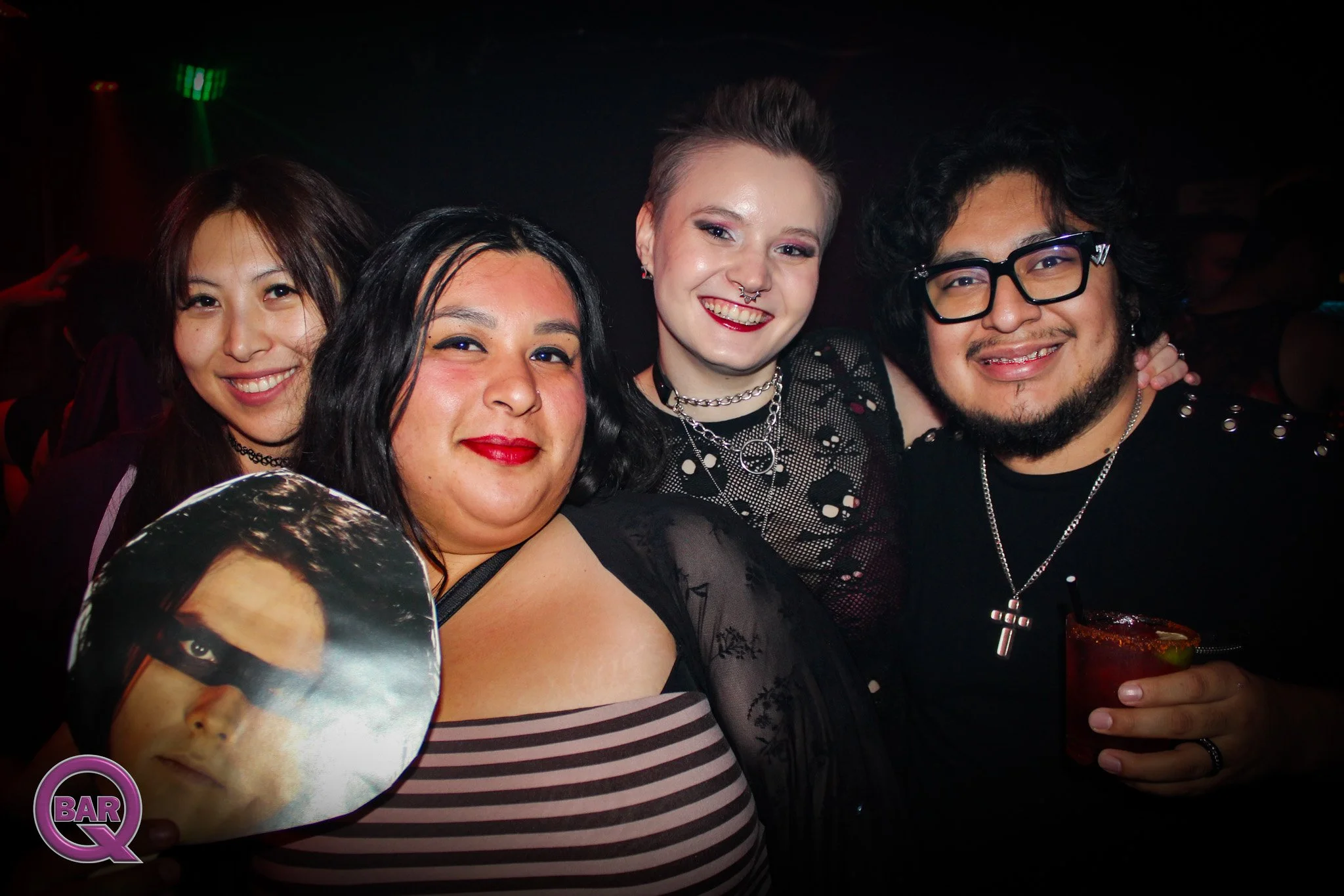 A group of four friends smiling at a party or nightclub, with dark background and colorful lighting. One person holds a round fan with a face on it, and another drinks a cocktail with a lime garnish.