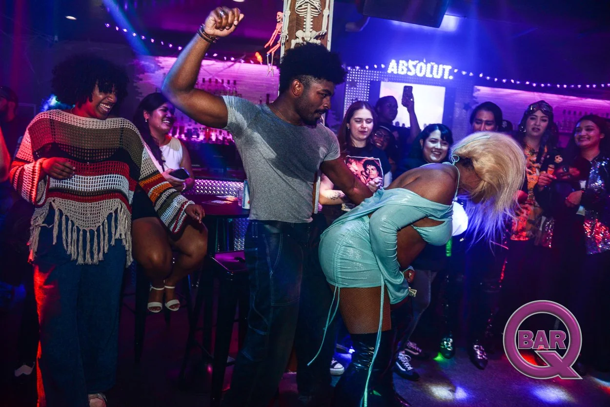 A lively nightclub scene with diverse people dancing, laughing, and enjoying music. A woman with blonde hair is bent over while a man holds her from behind. Colorful lights and a bar are visible in the background.