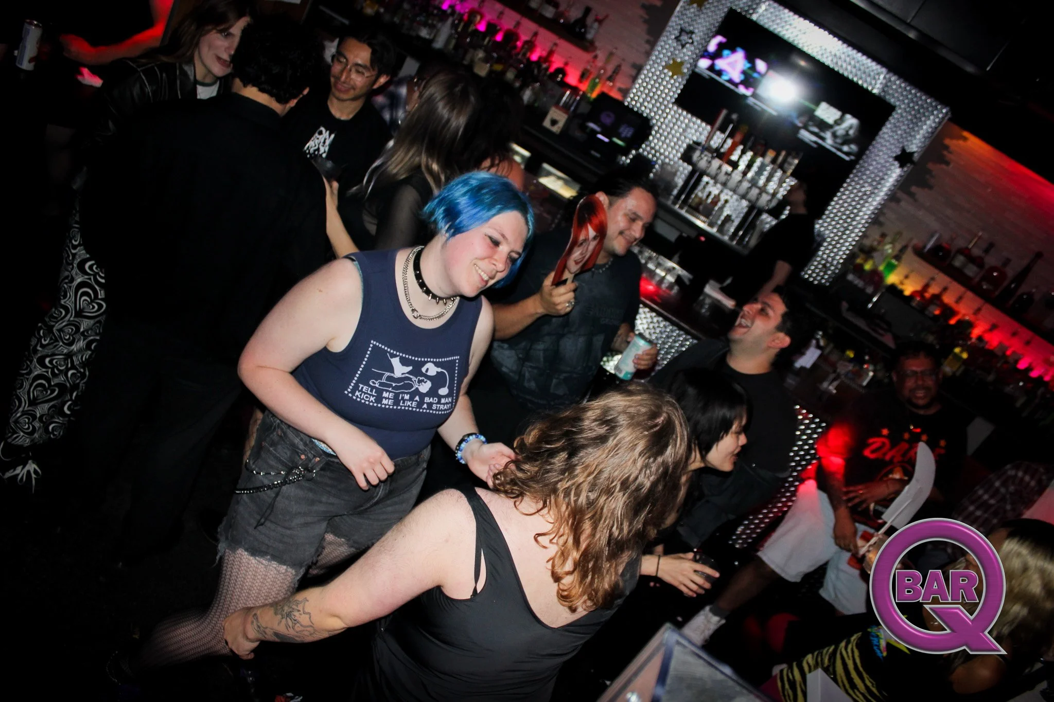 Group of people dancing and socializing in a dimly lit bar with shelves of liquor bottles and a neon sign in the background.