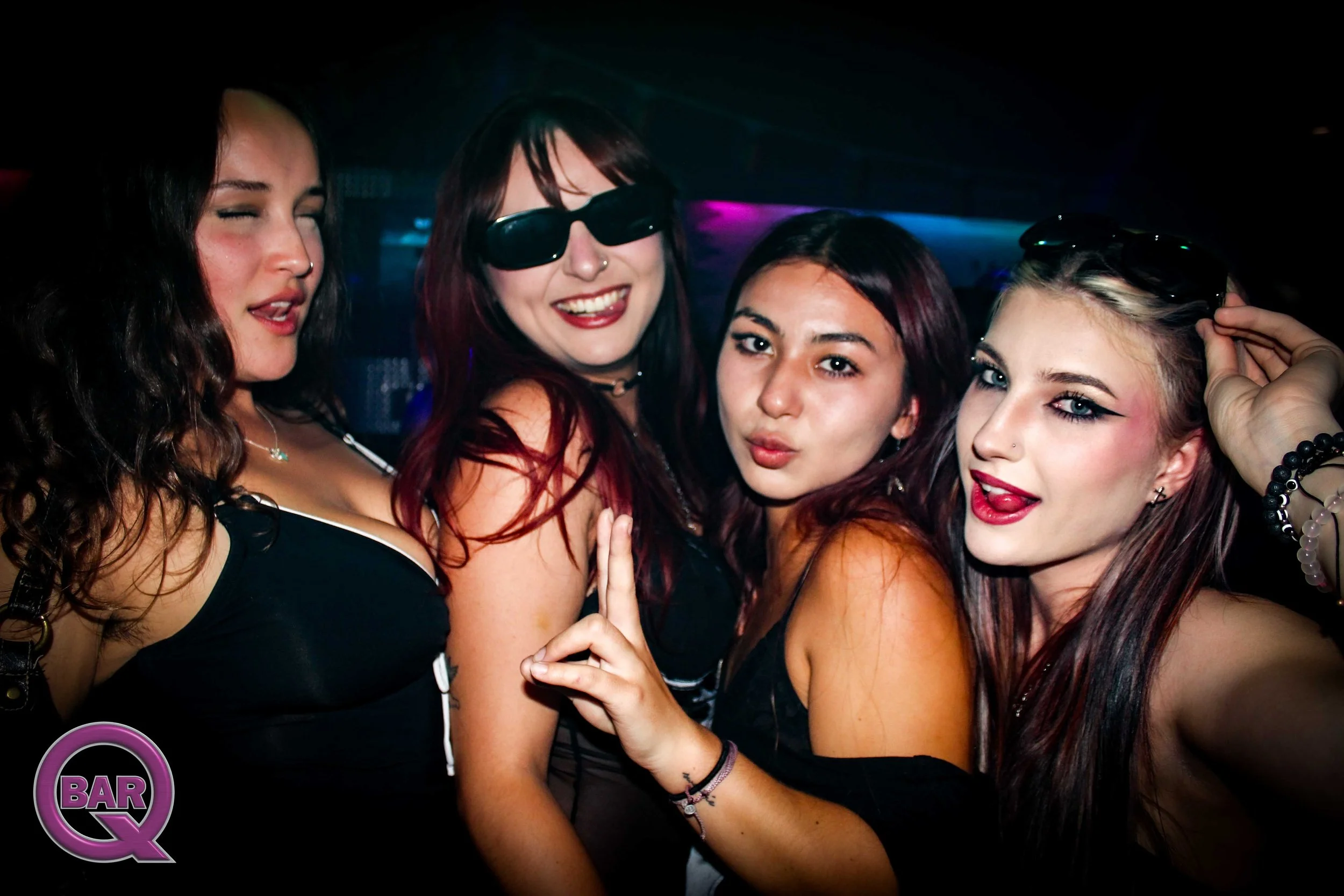 Four young women at a nightclub, smiling and posing for a photo, with dark background and neon lighting.