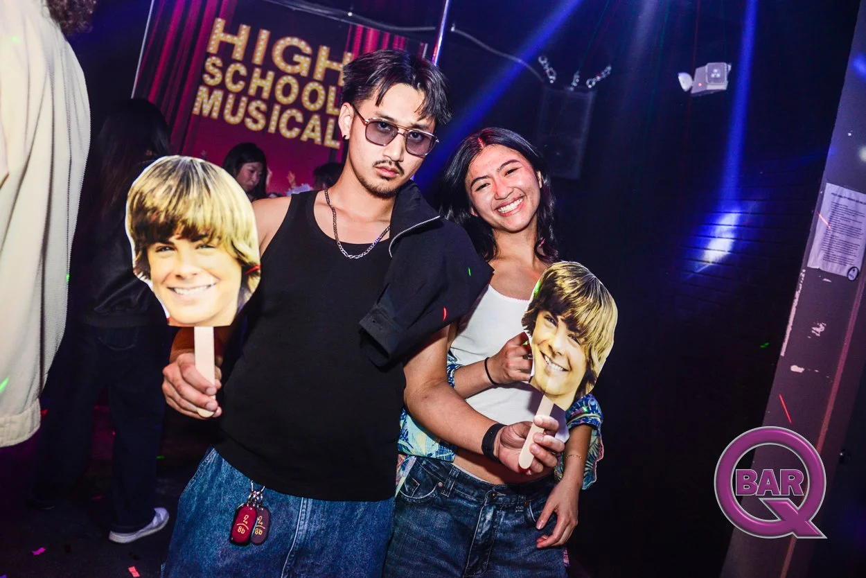 Two young people at a high school musical event holding cutouts of a young boy with blonde hair. The background has a sign that reads 'High School Musical' and a neon 'Bar Q' logo.