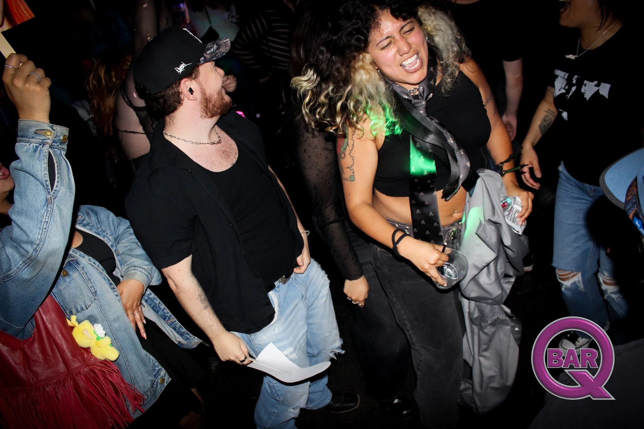 Group of young adults dancing and enjoying music at a bar or club, with a prominent smile on a woman with curly hair in the center.