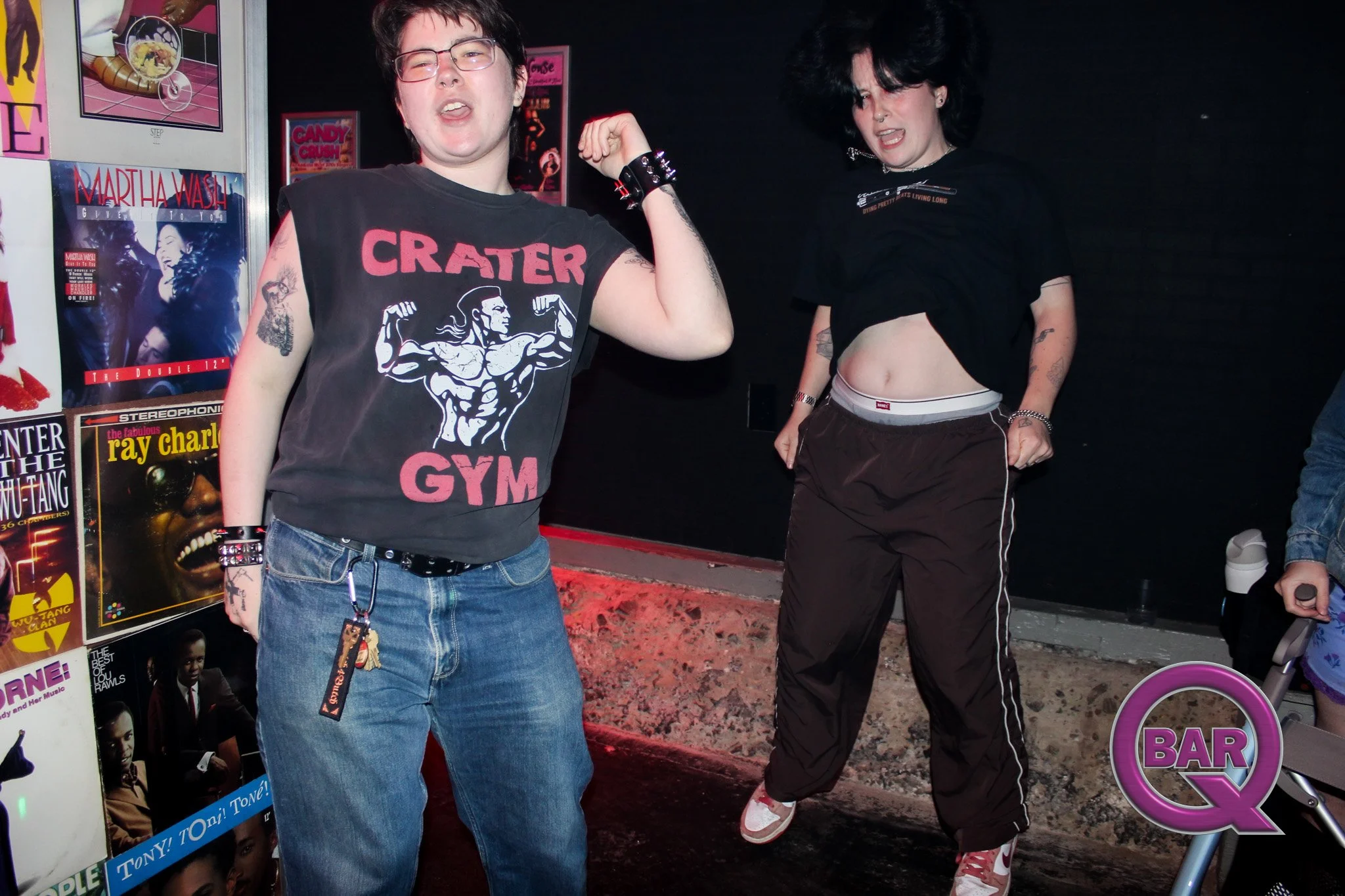 Two people with dark hair and tattoos, one wearing a black sleeveless shirt and the other lifting a black t-shirt, behind a wall of colorful posters and flyers, at a nightclub or bar.
