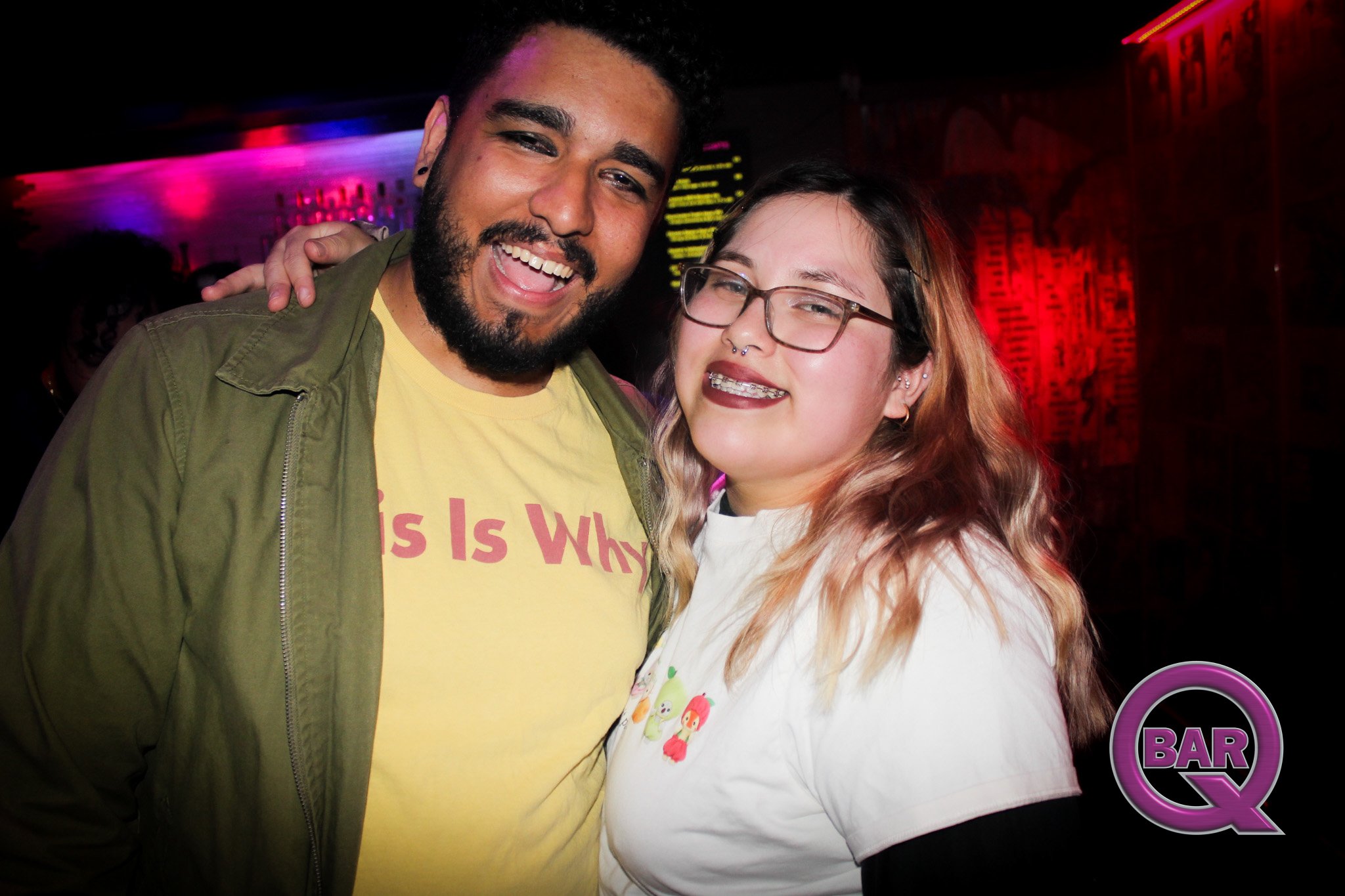 Two smiling people standing close together at a nightclub, with colorful lights in the background. The person on the left has dark hair, a beard, and is wearing a yellow shirt and an olive green jacket. The person on the right has light hair, glasses