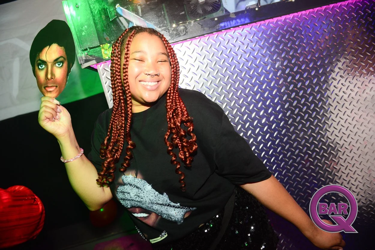 A smiling woman with long braided red hair and a nose ring holding a cardboard cutout of a woman with short black hair and makeup at a nightclub. The background has metallic diamond plate flooring, colorful lights, and a sign that says 'BAR Q'.