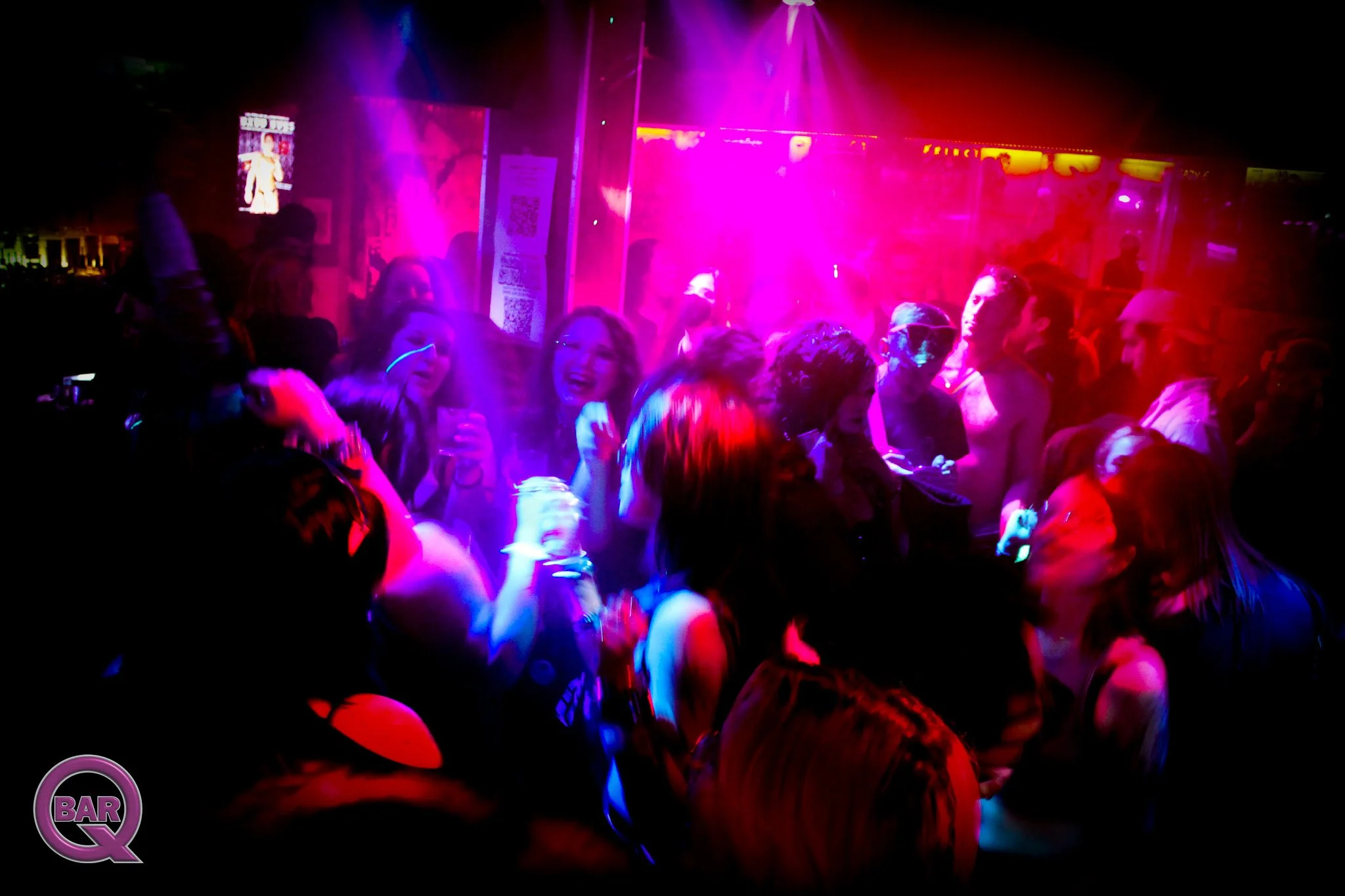 A crowded nightclub dance floor illuminated with pink, purple, and blue lights, with people dancing and socializing.