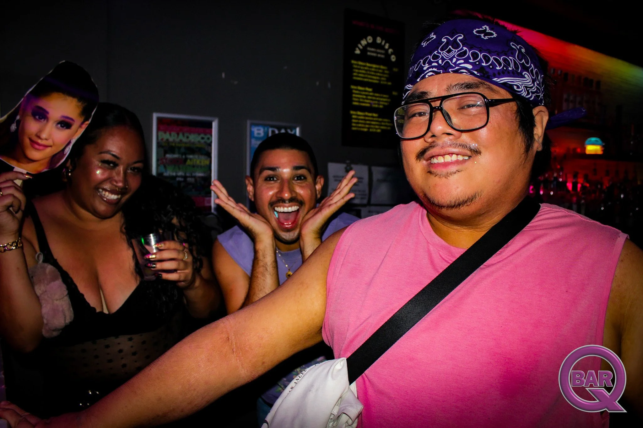 A group of four people at a bar or club, smiling and posing for the camera. The person in the foreground wears glasses, a bandana, and a pink sleeveless shirt, while the others include a woman holding a cutout of a woman's face and two men, one with 