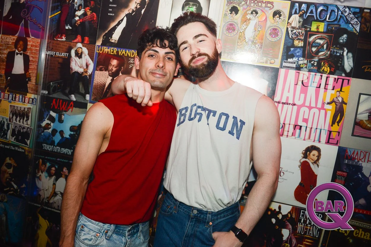 Two men standing close together with one man's arm around the other's shoulder in front of a wall decorated with album covers. The man on the left is wearing a sleeveless red shirt and has curly dark hair, while the man on the right is wearing a slee