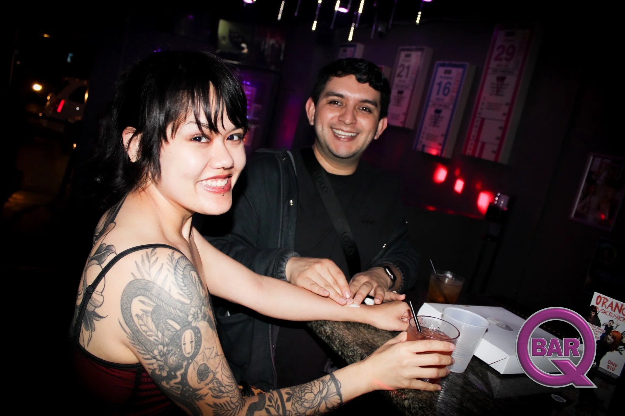 Two smiling people sitting at a bar counter with drinks, in a dimly lit bar or nightclub with posters on the wall.