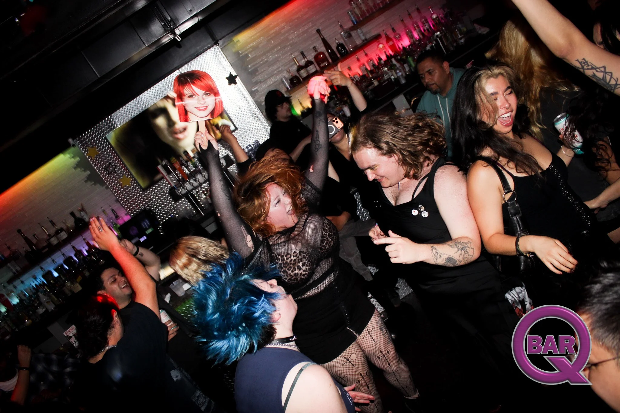 A lively bar scene with multiple women dancing and socializing. One woman with bright red hair holds up a paper fan with a woman's face on it, others have colorful hair and tattoos. The bar is stocked with bottles and illuminated with colorful lights