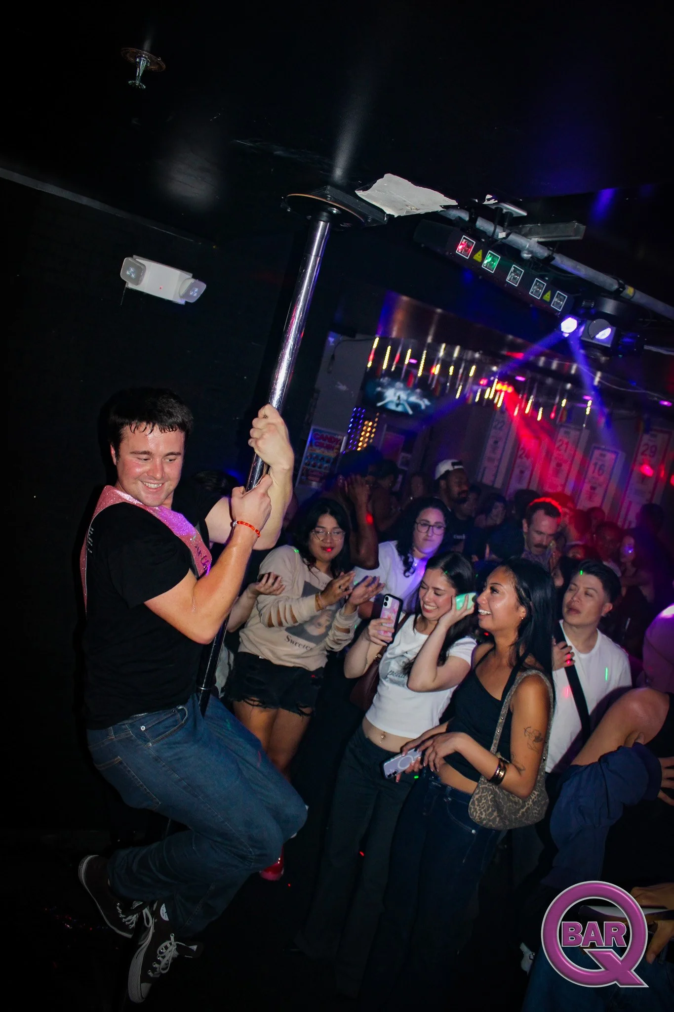 A man dancing on a pole in a crowded bar with people taking photos and enjoying the lively atmosphere.