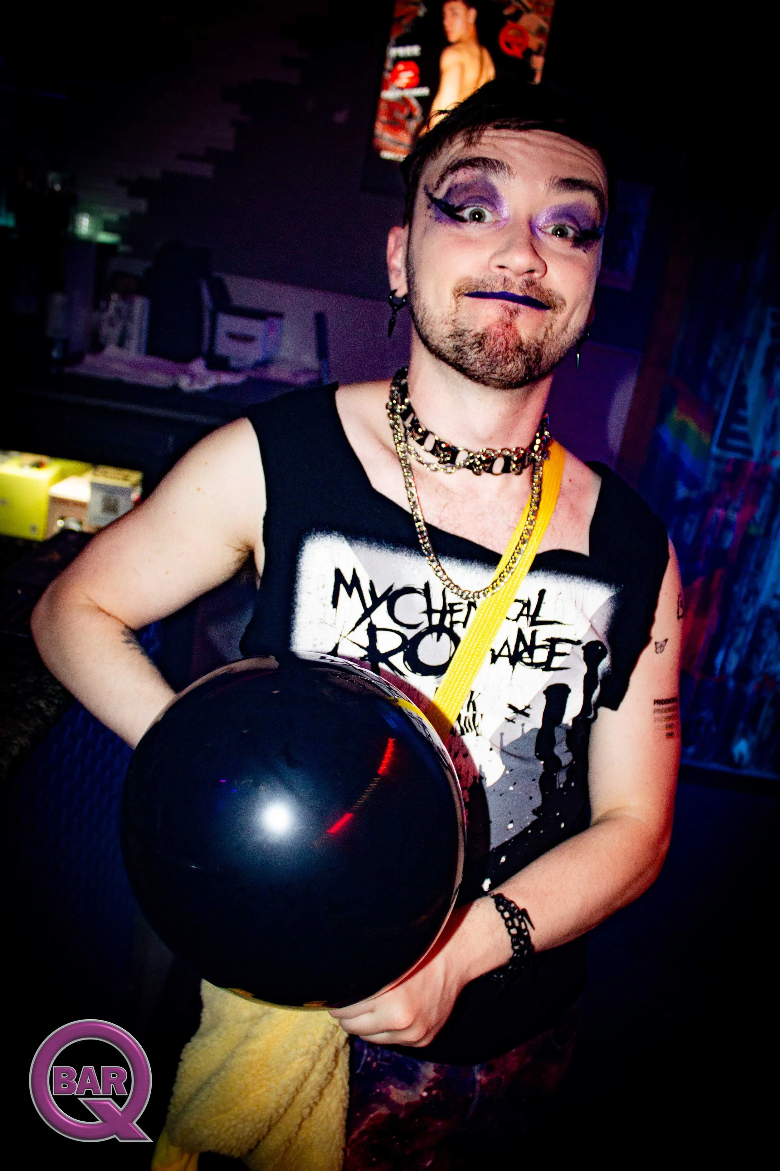 Person with colorful eye makeup, wearing a black sleeveless shirt with 'My Chemical Romance' text, holding a black balloon, and standing in a dimly lit room with posters and colorful decorations.
