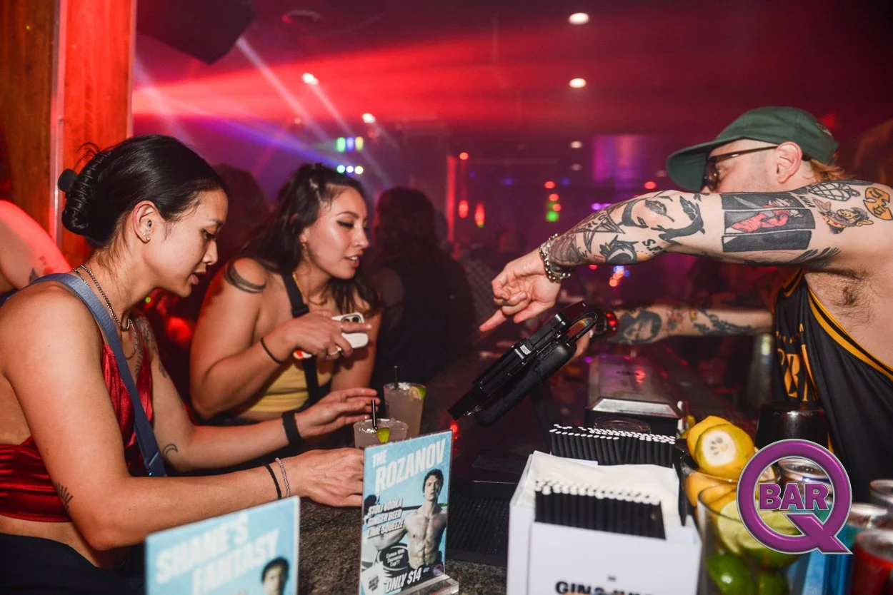 Women at a bar ordering drinks from a tattooed bartender with fluorescent lighting and party atmosphere in background.