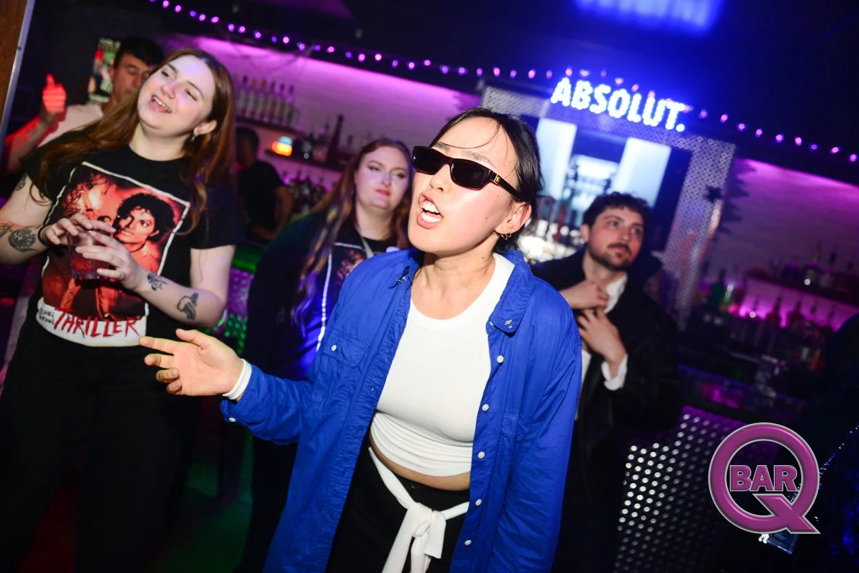 Group of people dancing and socializing at a bar or nightclub with colourful lighting and the word 'ABSOLUT' illuminated in the background.