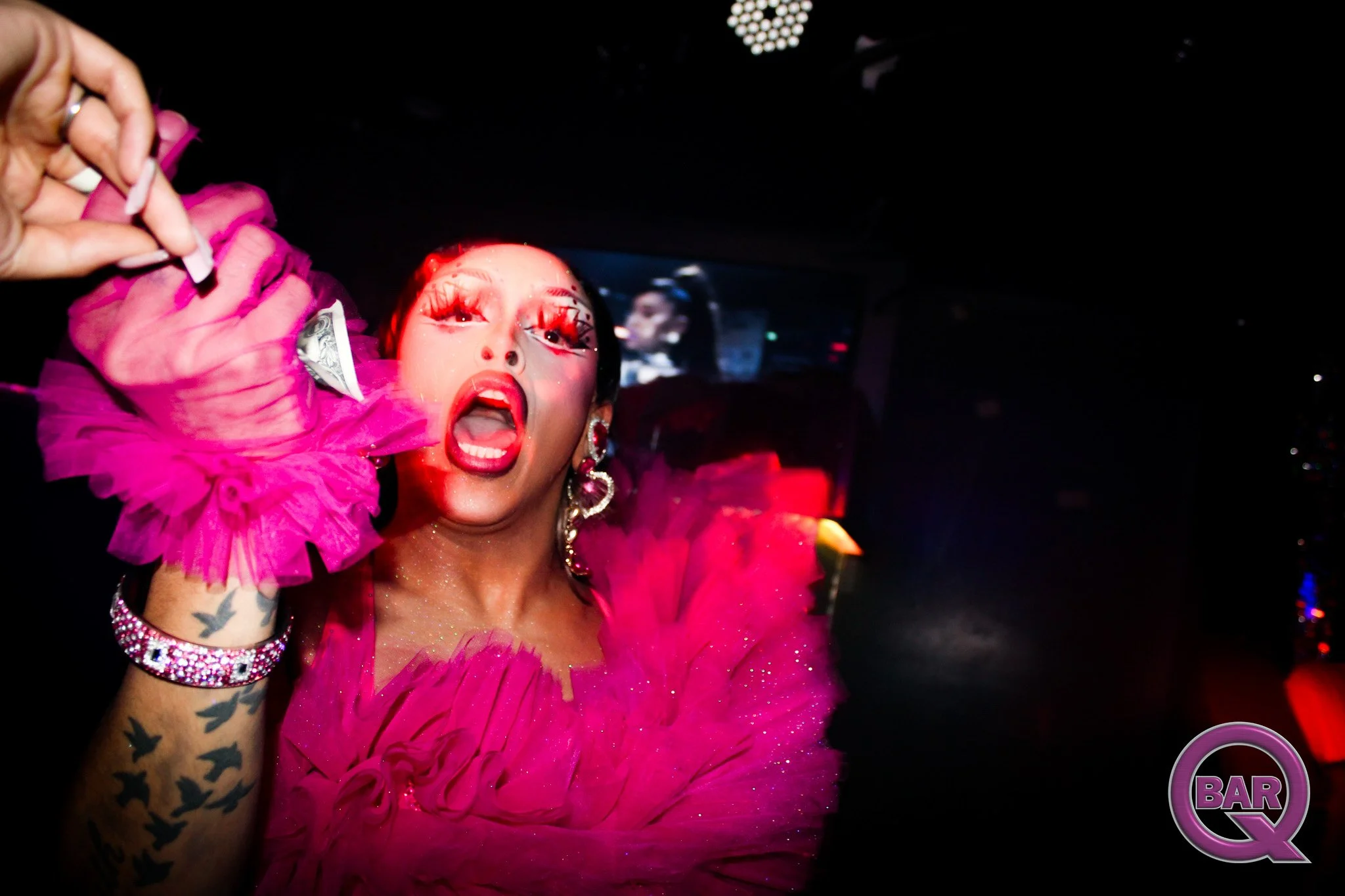 A woman in a bright pink, ruffled dress with large earrings and a bracelet, with her mouth open and eyes wide, inside a dark venue with a screen in the background showing her reflection.