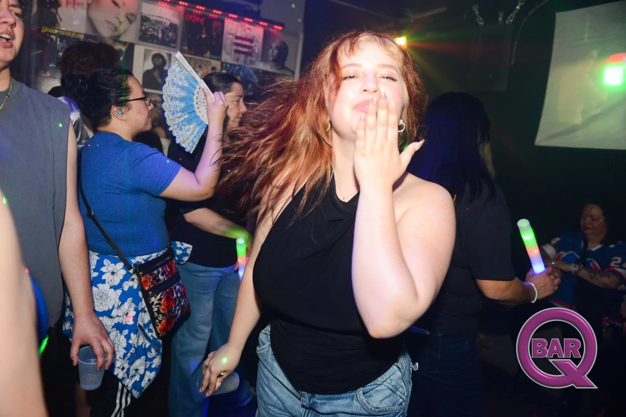 A woman with red hair blowing a kiss at a lively dance party, surrounded by other partygoers in a club with record covers on the wall and colorful lighting.