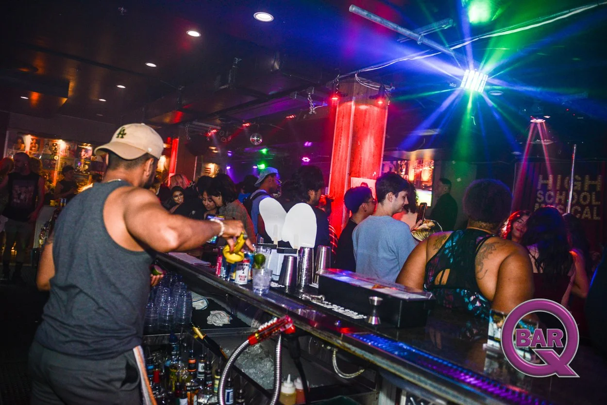 Crowded nightclub with people dancing and socializing, illuminated by colorful disco lights, with a bartender pouring a drink in the foreground.