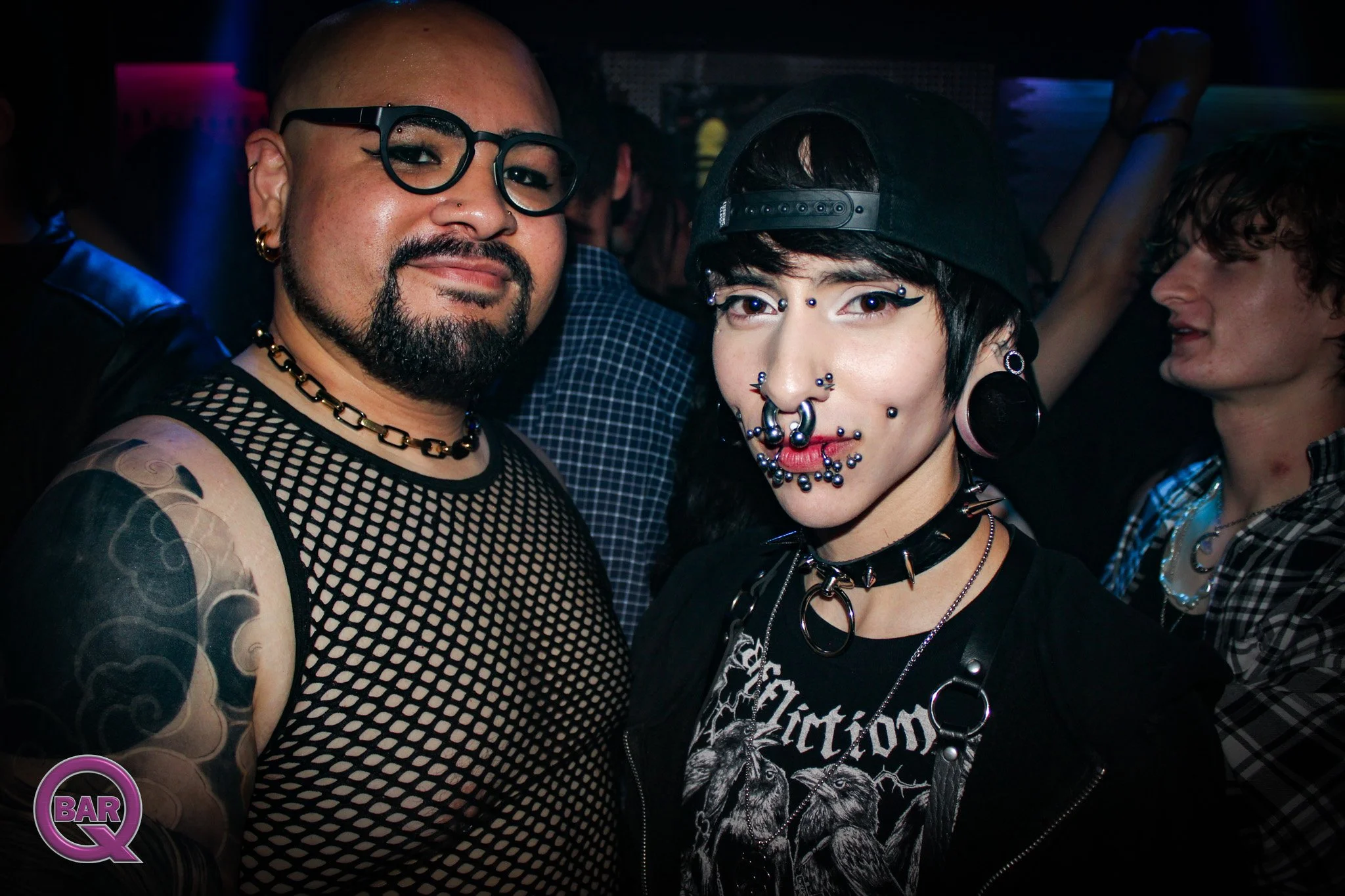 Two people at a party or club, one with glasses and a mesh shirt, the other with piercings, jewelry, and a black cap, labeled as 'Bar Q'.
