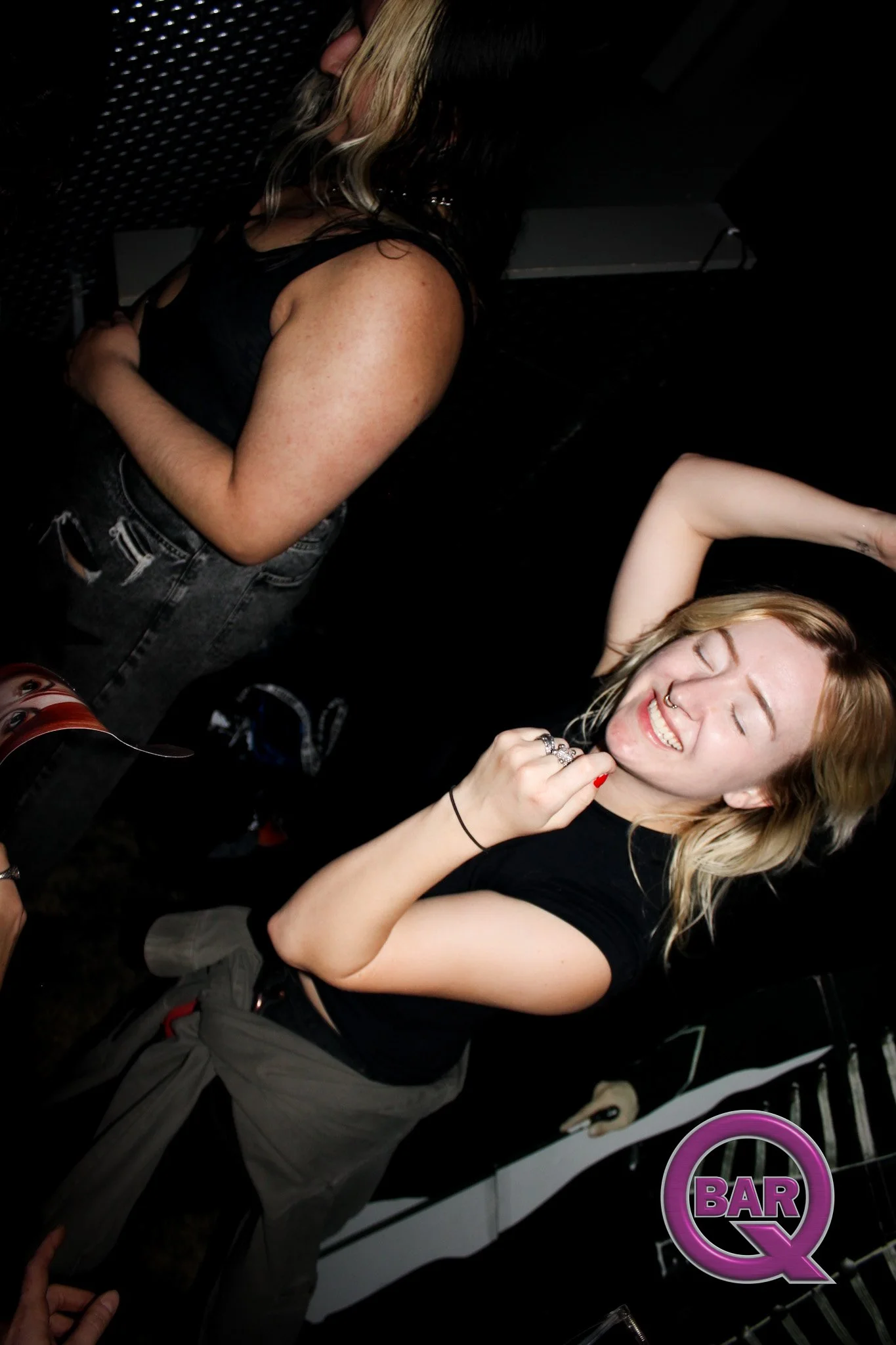 A young woman with blonde hair smiling with eyes closed at a lively bar or nightclub. She is wearing a black T-shirt and has multiple rings on her fingers. The background features a dark venue with people and a neon sign that says "BAR Q".
