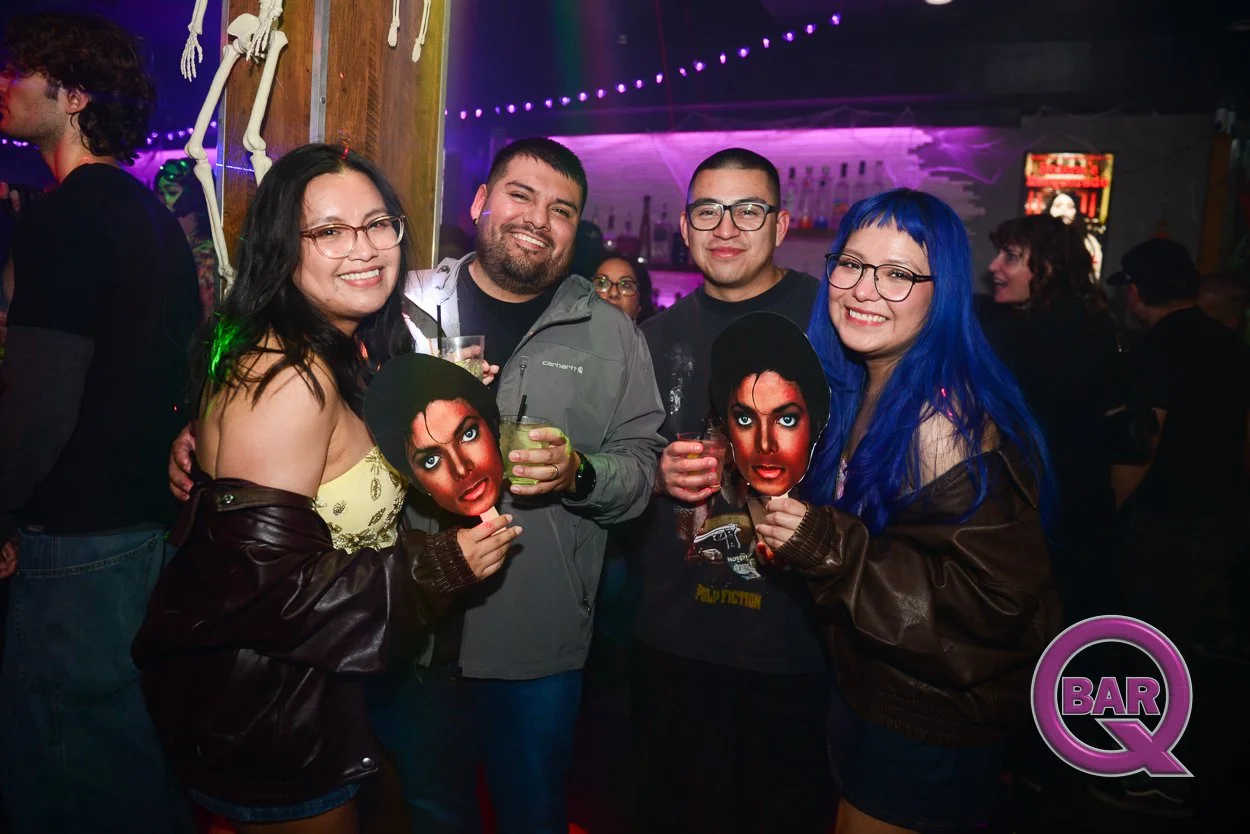 Group of four friends smiling and posing with Michael Jackson masks at a bar or nightclub, with neon lights and other patrons in the background.
