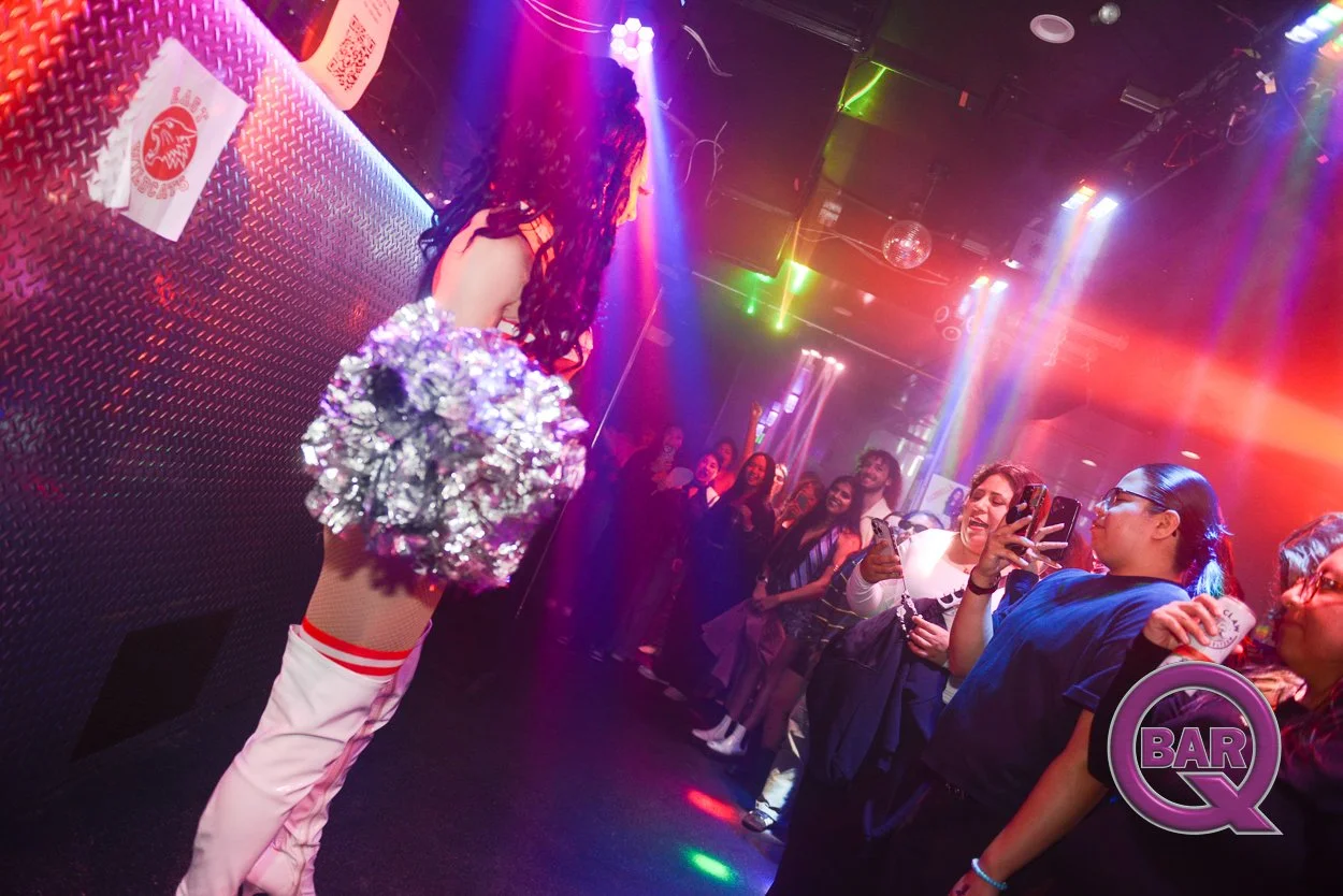 A performer in a nightclub, dressed in a sporty outfit with shiny pom-poms, faces an audience of excited women taking photos and videos with their phones under colorful lights.