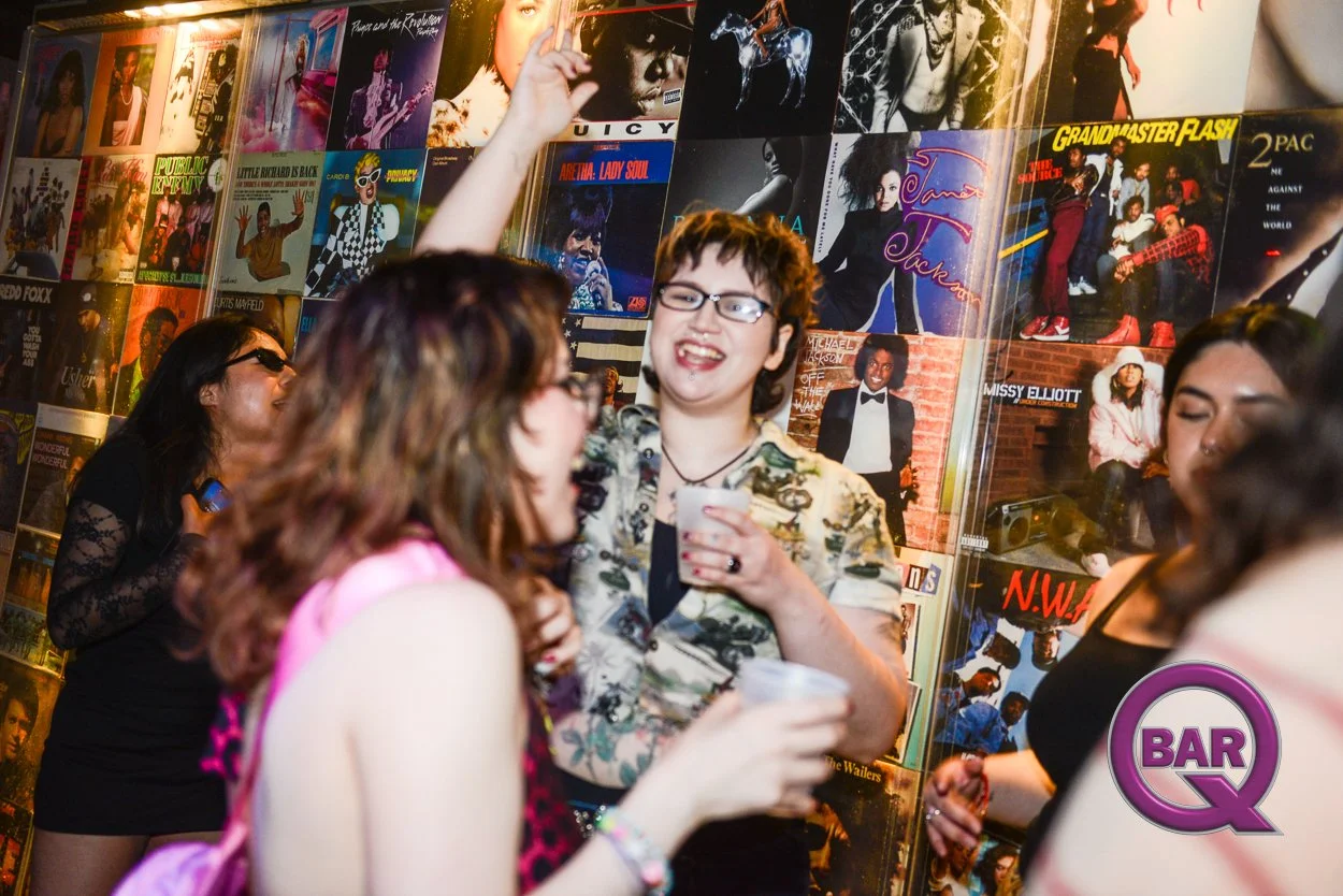 A group of people socializing at a bar, with a colorful wall of music album covers in the background.