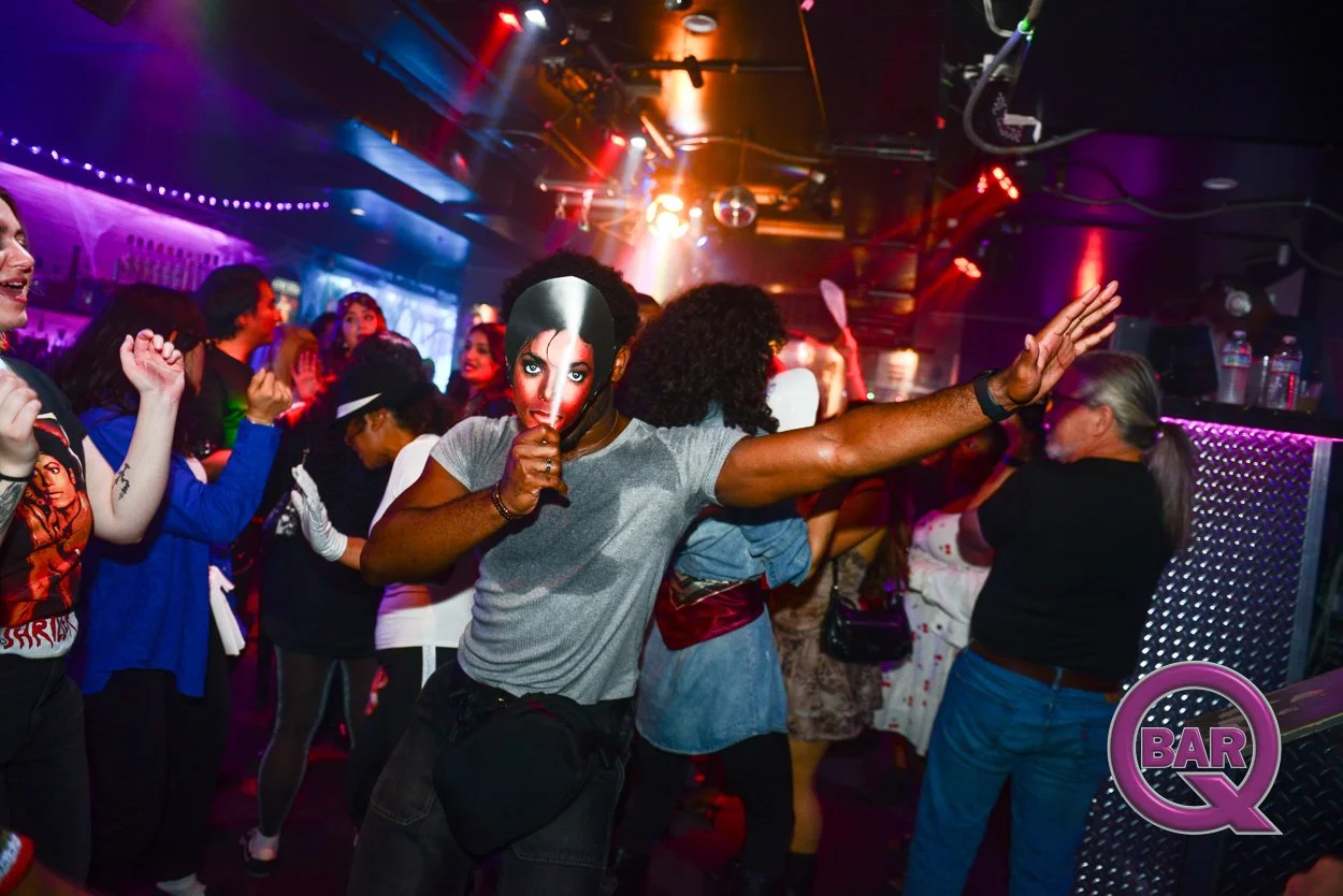People dancing at a nightclub, some wearing masks, with colorful lights and a bar in the background.