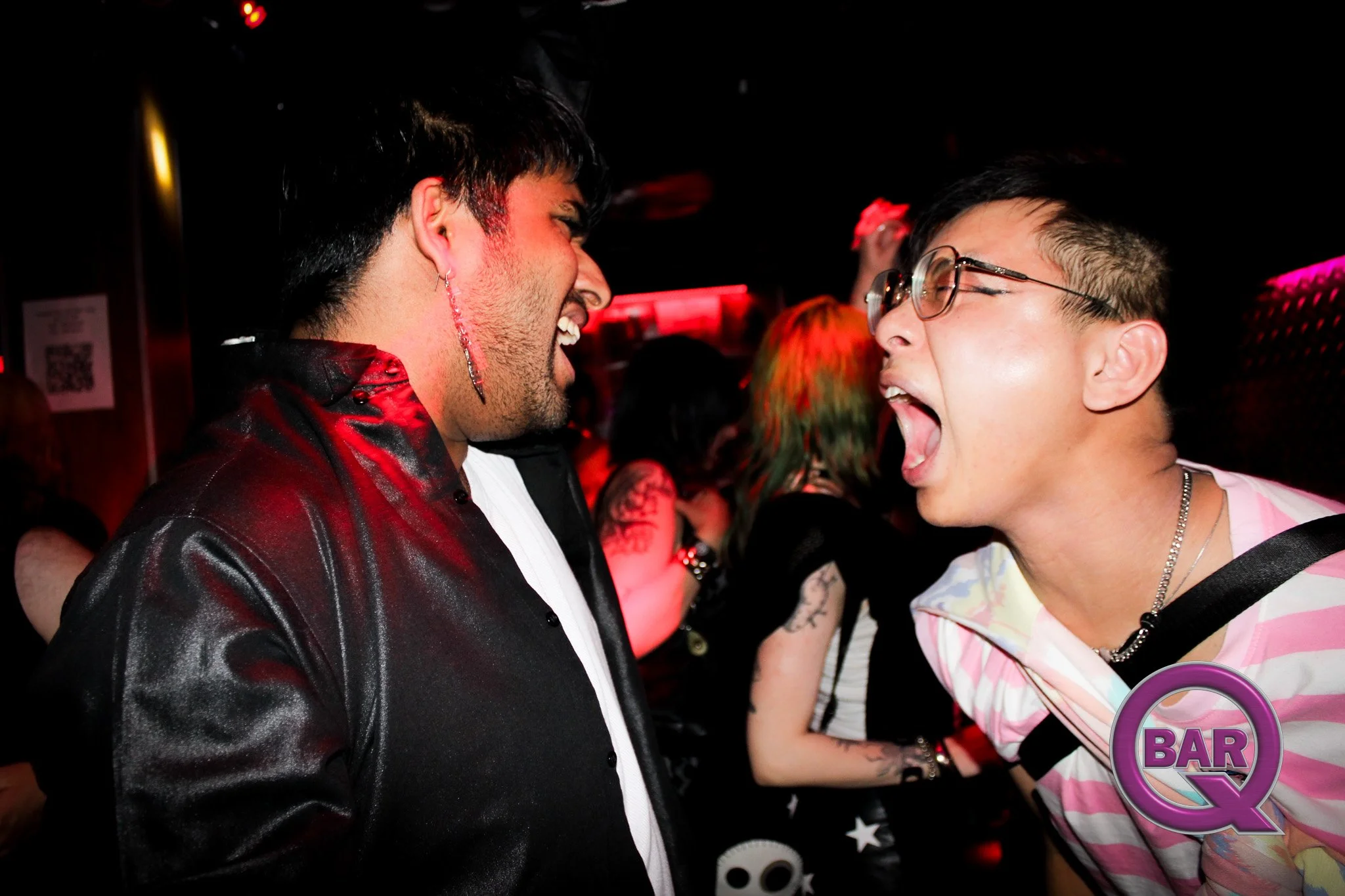 Two people facing each other and smiling, enjoying a moment of laughter in a dimly lit bar or nightclub with red and pink lighting.