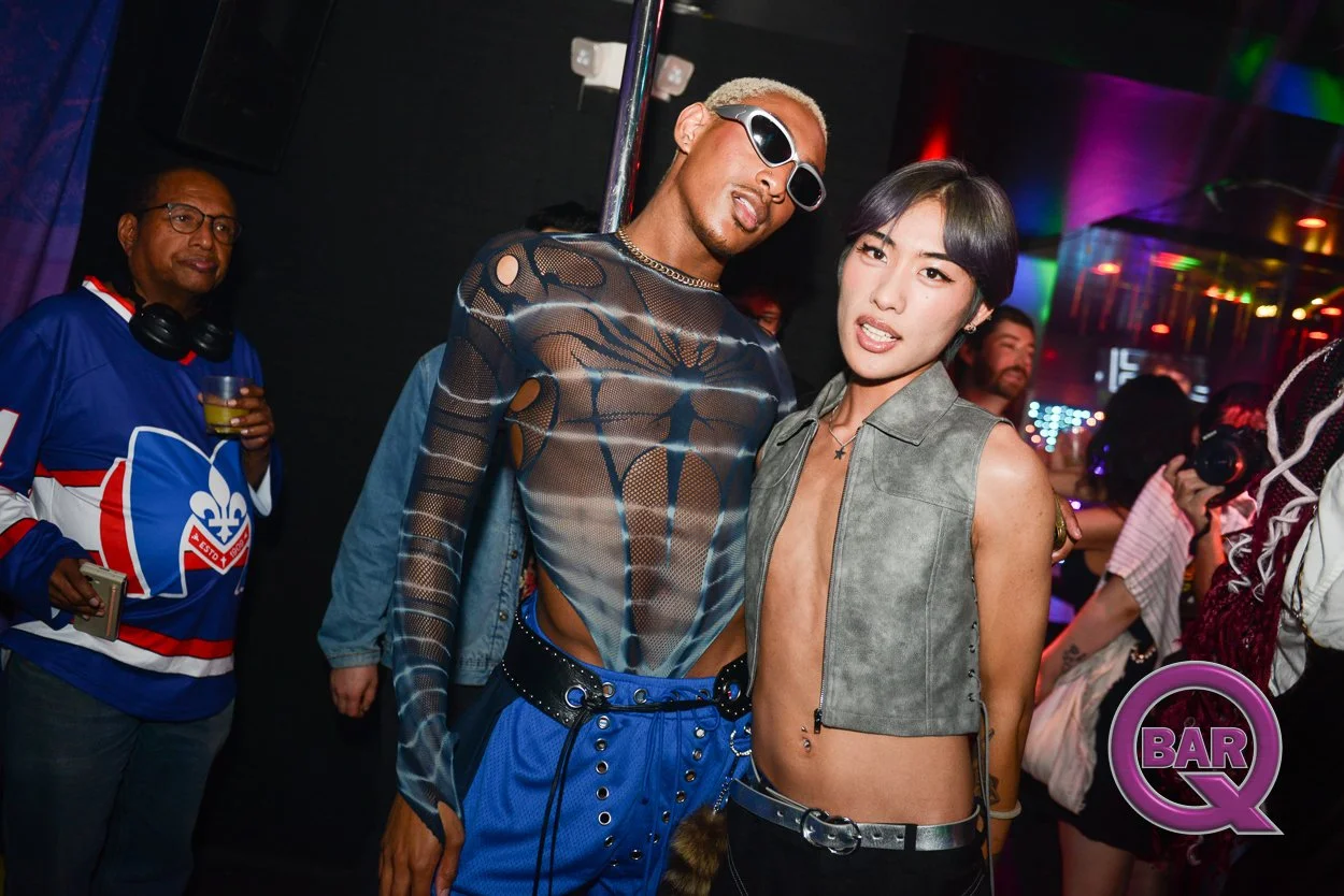 Two people posing at a nightclub, one wearing sunglasses and a sheer top, the other in a sleeveless gray shirt with a deep neckline, surrounded by other partygoers and colorful lights.