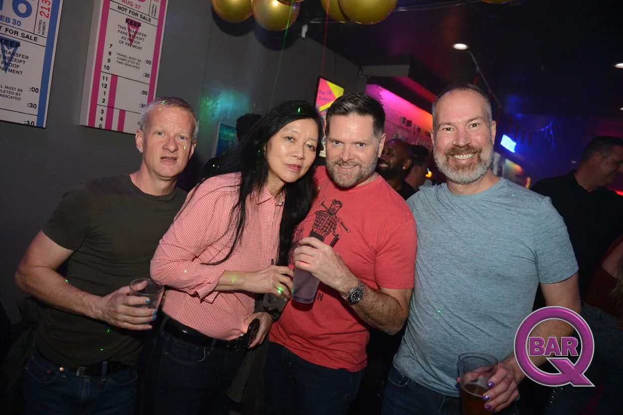 Four people smiling and holding drinks at a bar or nightclub, with colorful lighting, balloons, and posters on the wall.
