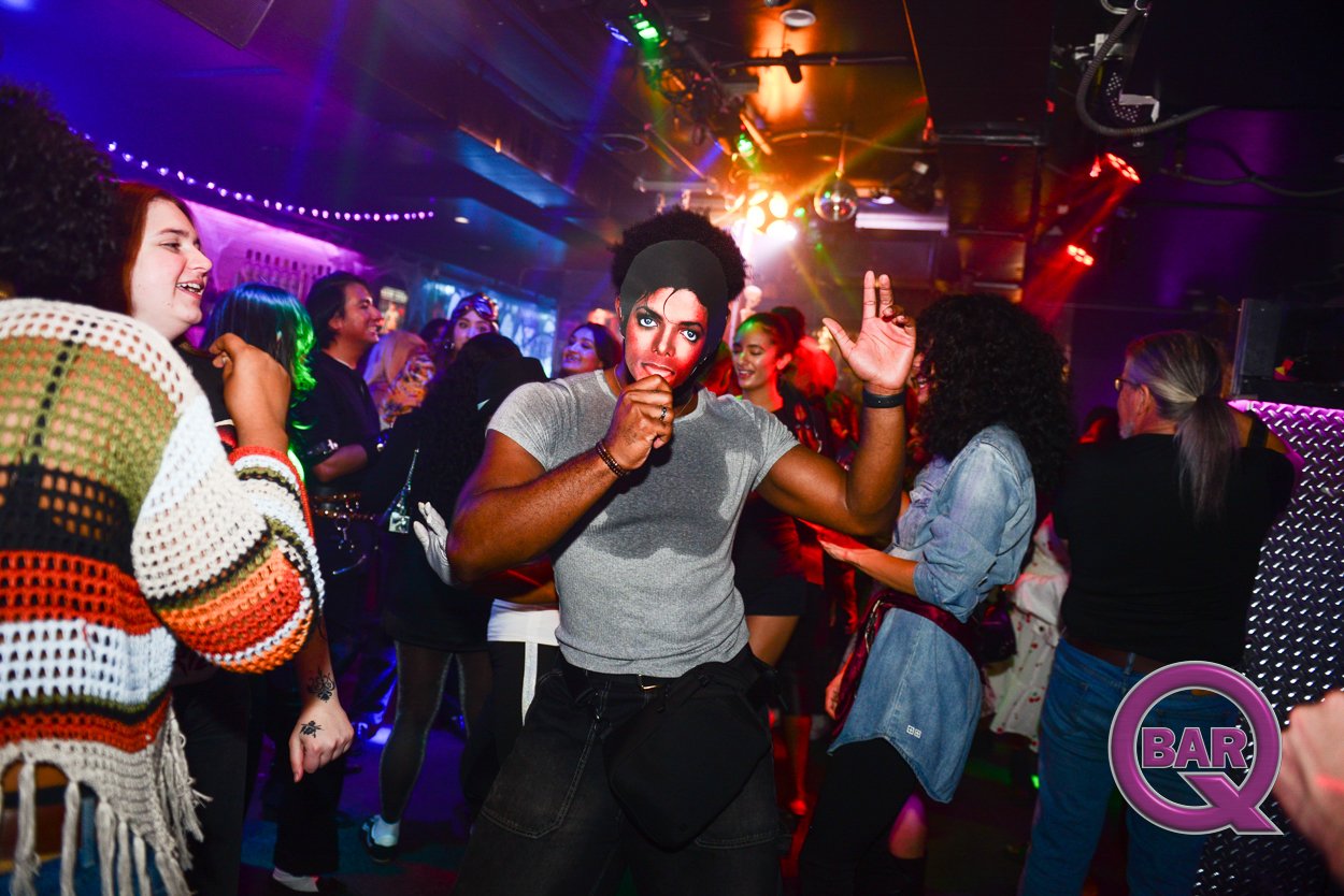 People dancing and socializing in a nightclub with colorful lights and a DJ booth in the background.