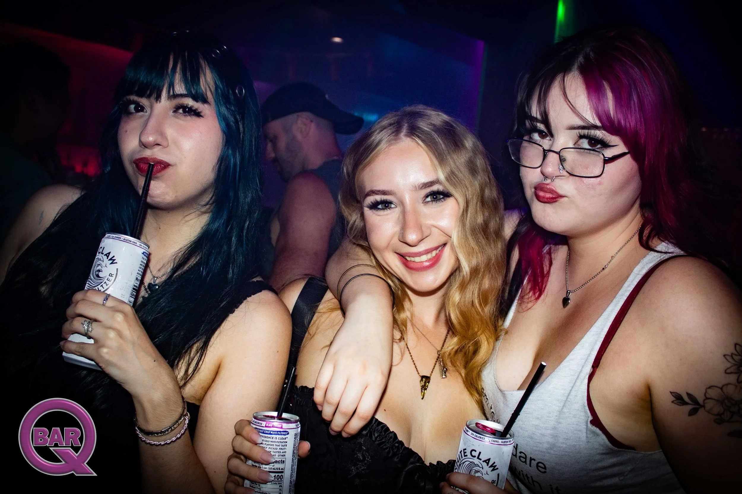 Three young women at a nightclub or party holding cans of drinks, smiling, with colorful lights and a dark background.