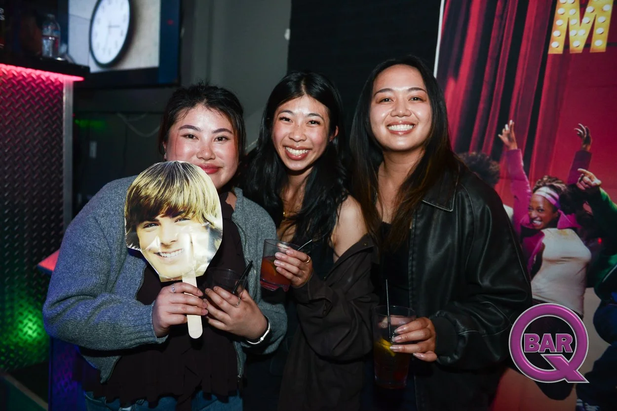 Three women at a bar or club, smiling and holding drinks; one woman holds a Jack Nicholson mask. A vibrant backdrop with people dancing and a bar logo in the corner.